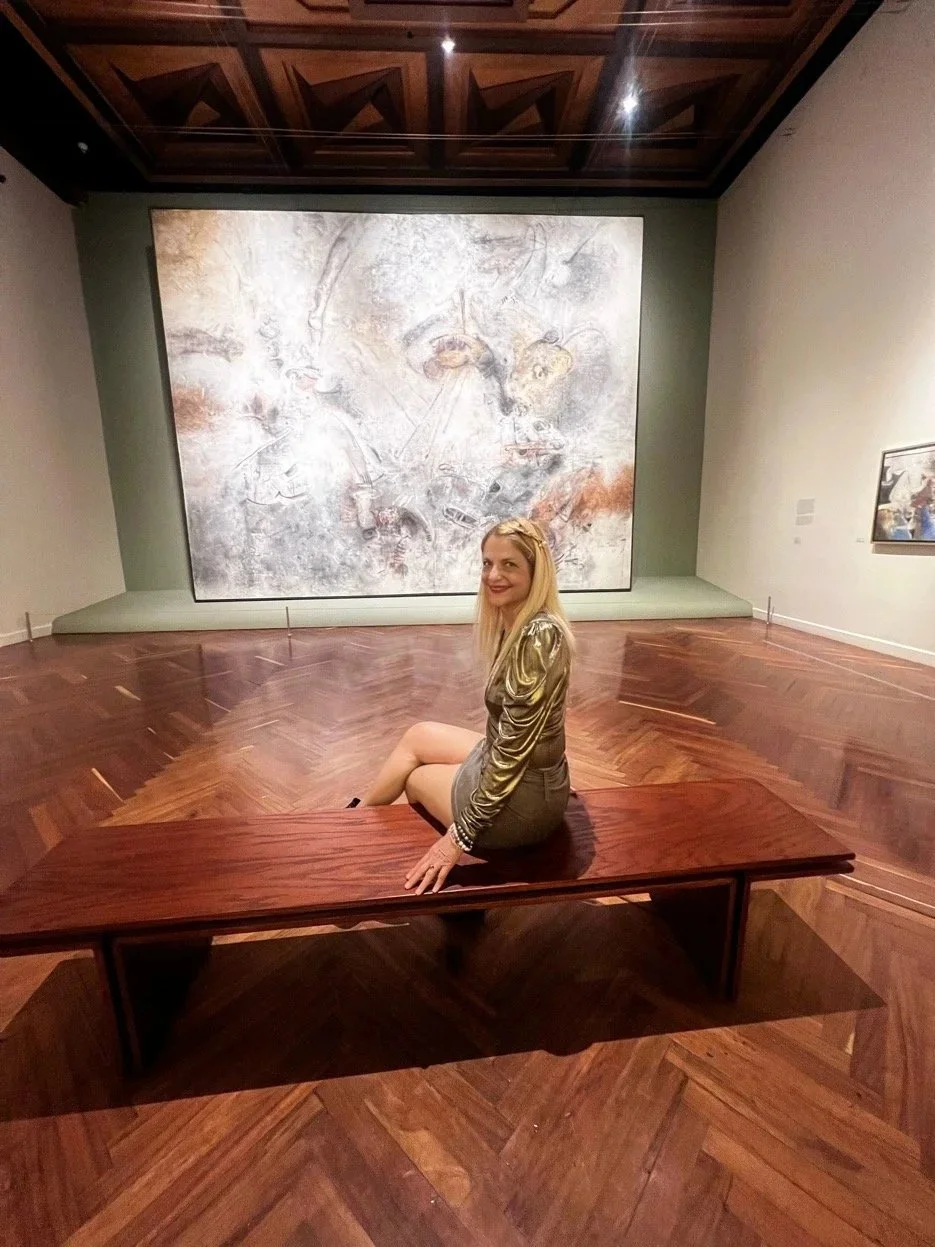 A woman with long blonde hair sitting on a wooden bench in an art gallery, smiling at the camera. Behind her is a large abstract painting on the wall, and the gallery has wooden floors and a wooden ceiling.