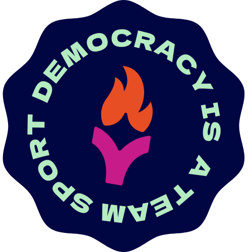 A circular badge with the words 'DEMOCRACY IS TEAM SPORT' around a stylized flame symbol, featuring a purple hand and orange flames on a black background.