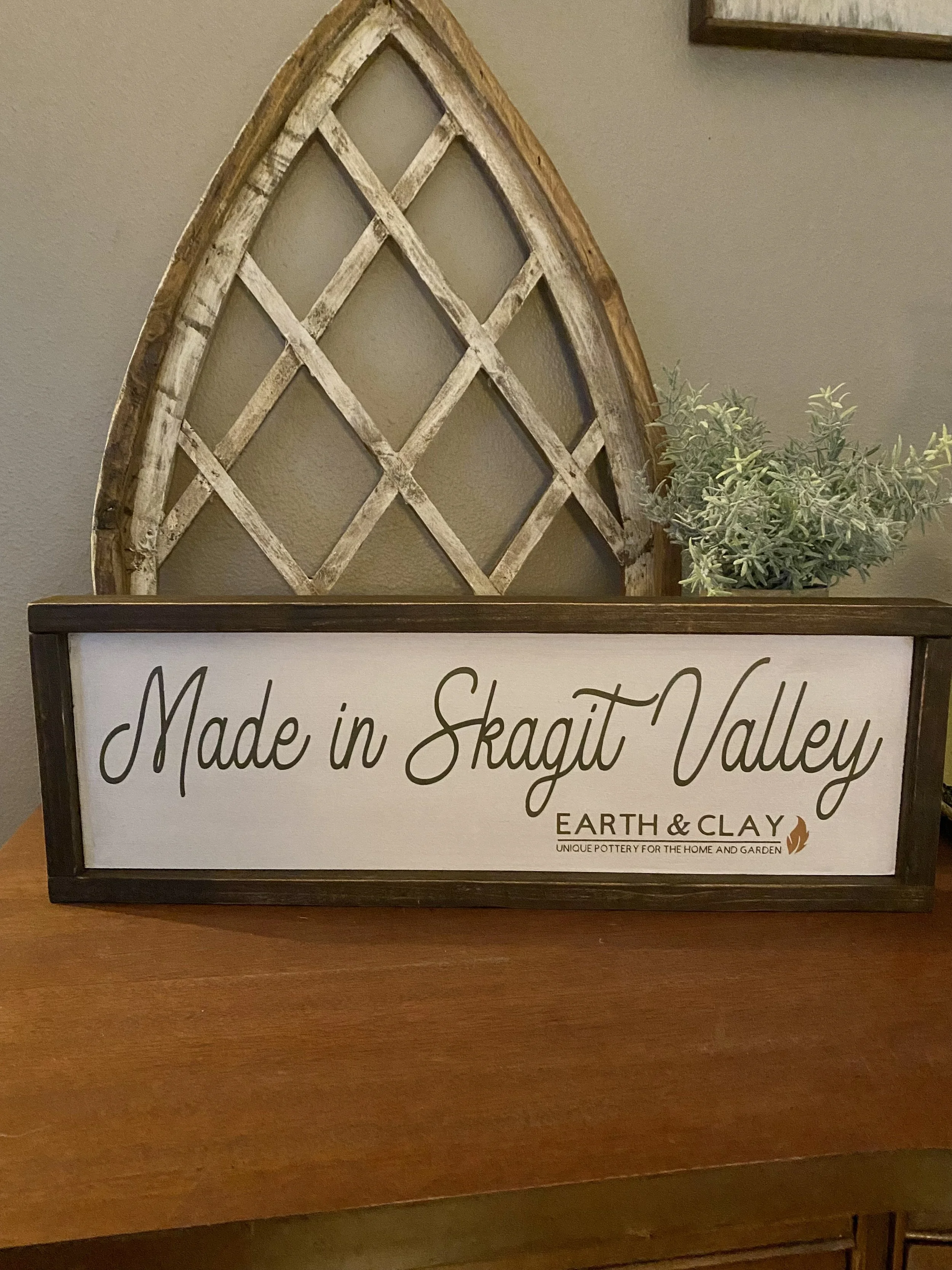 A decorative sign on a wooden surface reading 'Made in Skagit Valley' with a smaller text underneath that says 'Earth & Clay, Unique Pottery for the Home and Garden.' Behind the sign, there is a vintage wooden window frame leaning against the wall wi