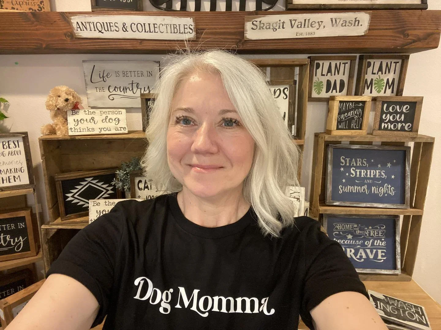 A woman with silver hair smiling in front of decorative signs and frames with quotes and phrases on a wooden display shelf.