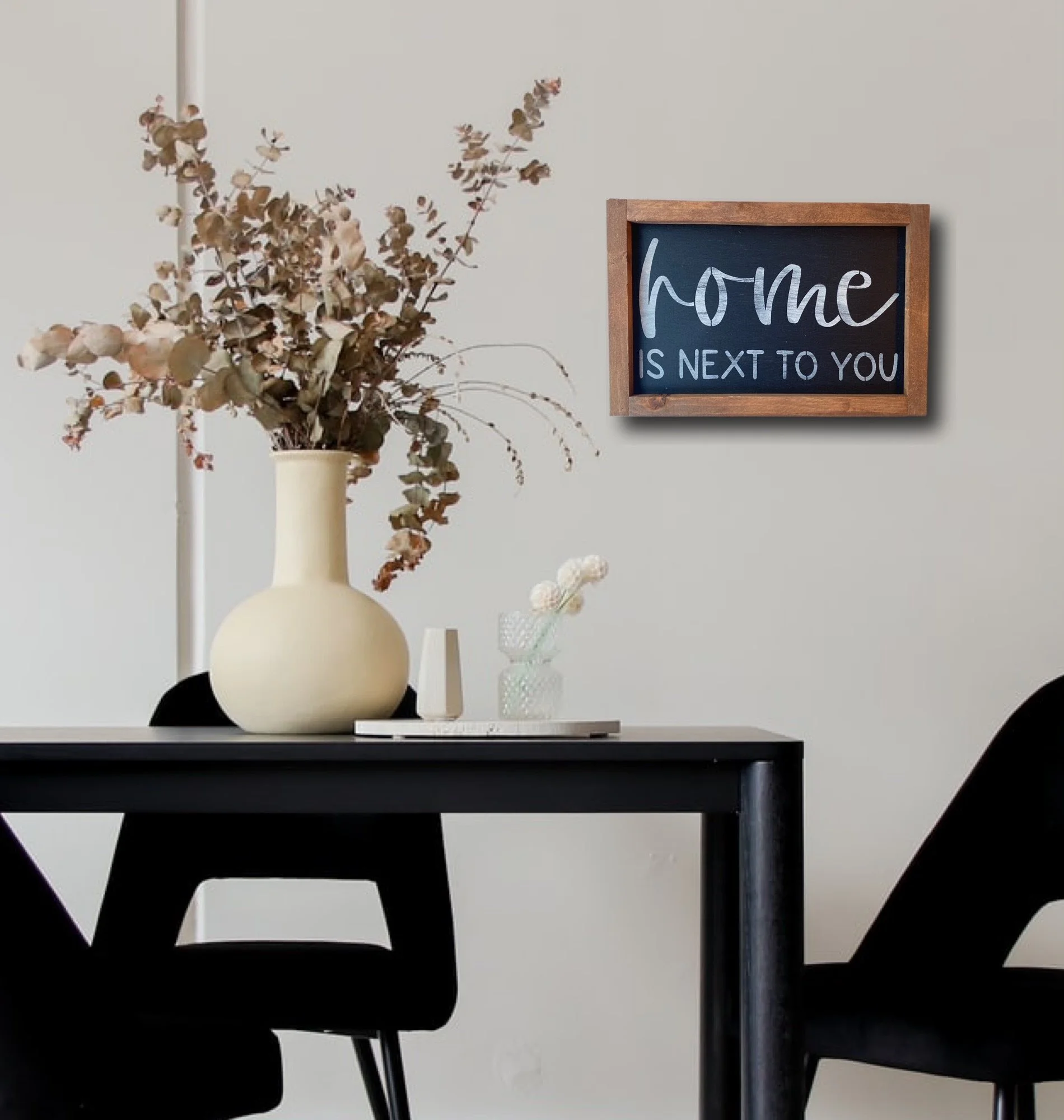 Decorative dining room with a black table, black chairs, a beige vase with dried flowers, a smaller glass vase, and a framed picture on the wall that reads "home is next to you".