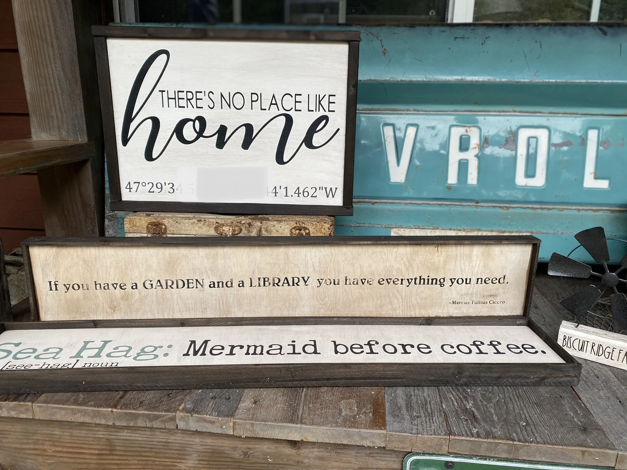 Three decorative signs with inspirational quotes, placed on a wooden surface outdoors. The top sign says, "There's no place like home" with coordinates. The middle sign quotes Marcus Tullius Cicero about having a garden and a library. The bottom sign