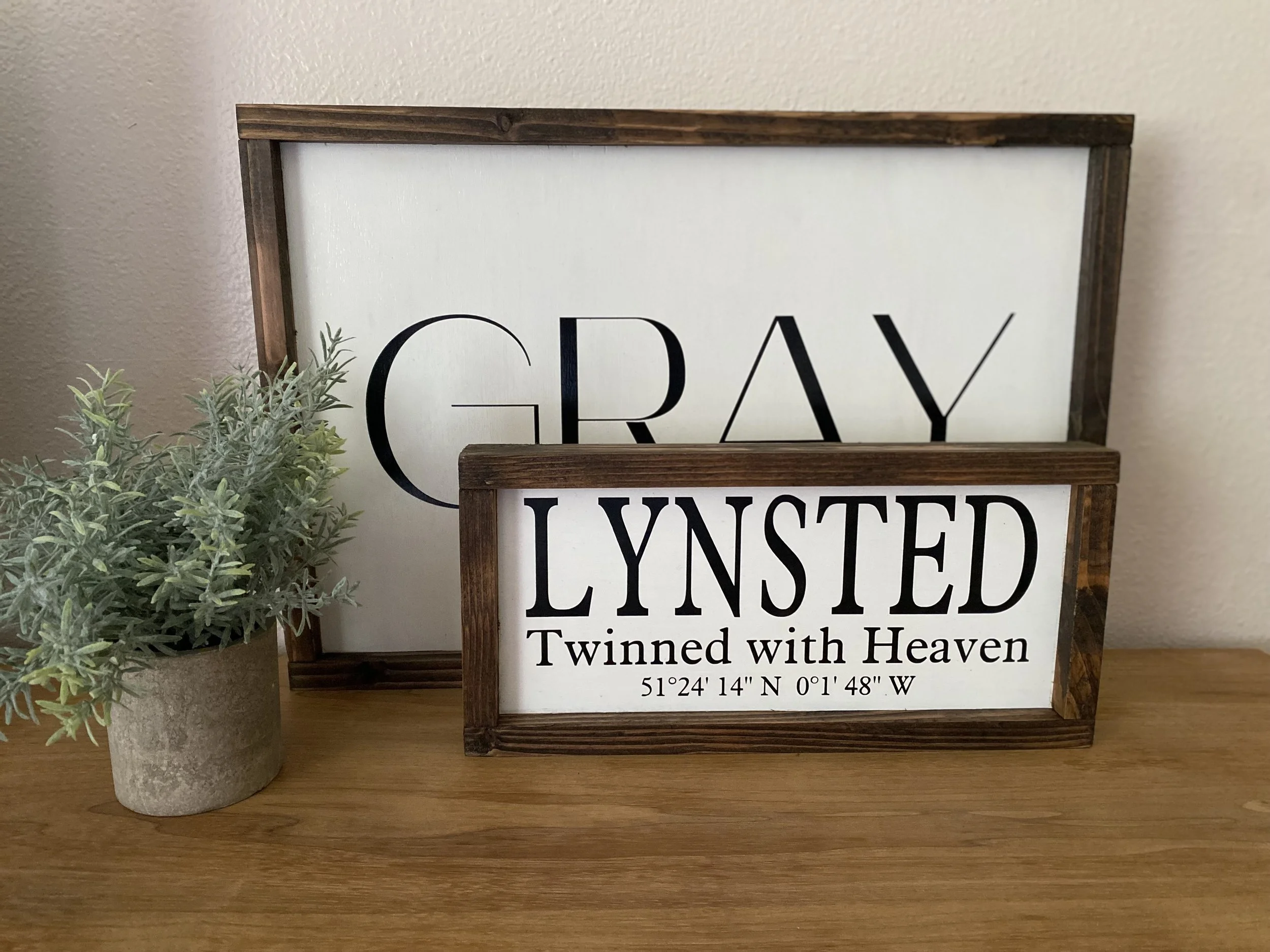 Decorative sign with the words 'GRAY LYNSTED' and 'Twinned with Heaven' along with geographical coordinates, placed on a wooden surface next to a small potted green plant.