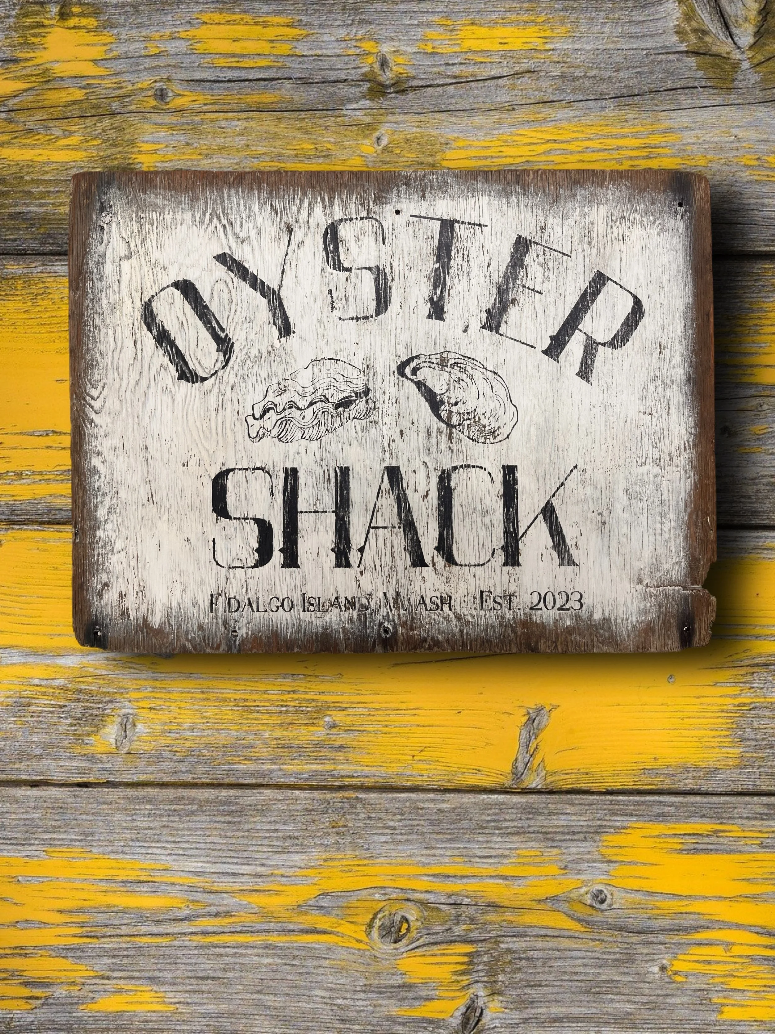 Wooden sign with black lettering that reads 'OYSTER SHACK' and 'FIDALGO ISLAND WASH EST. 2023', featuring illustrations of a clam and a crab, mounted on a weathered yellow and gray wooden surface.