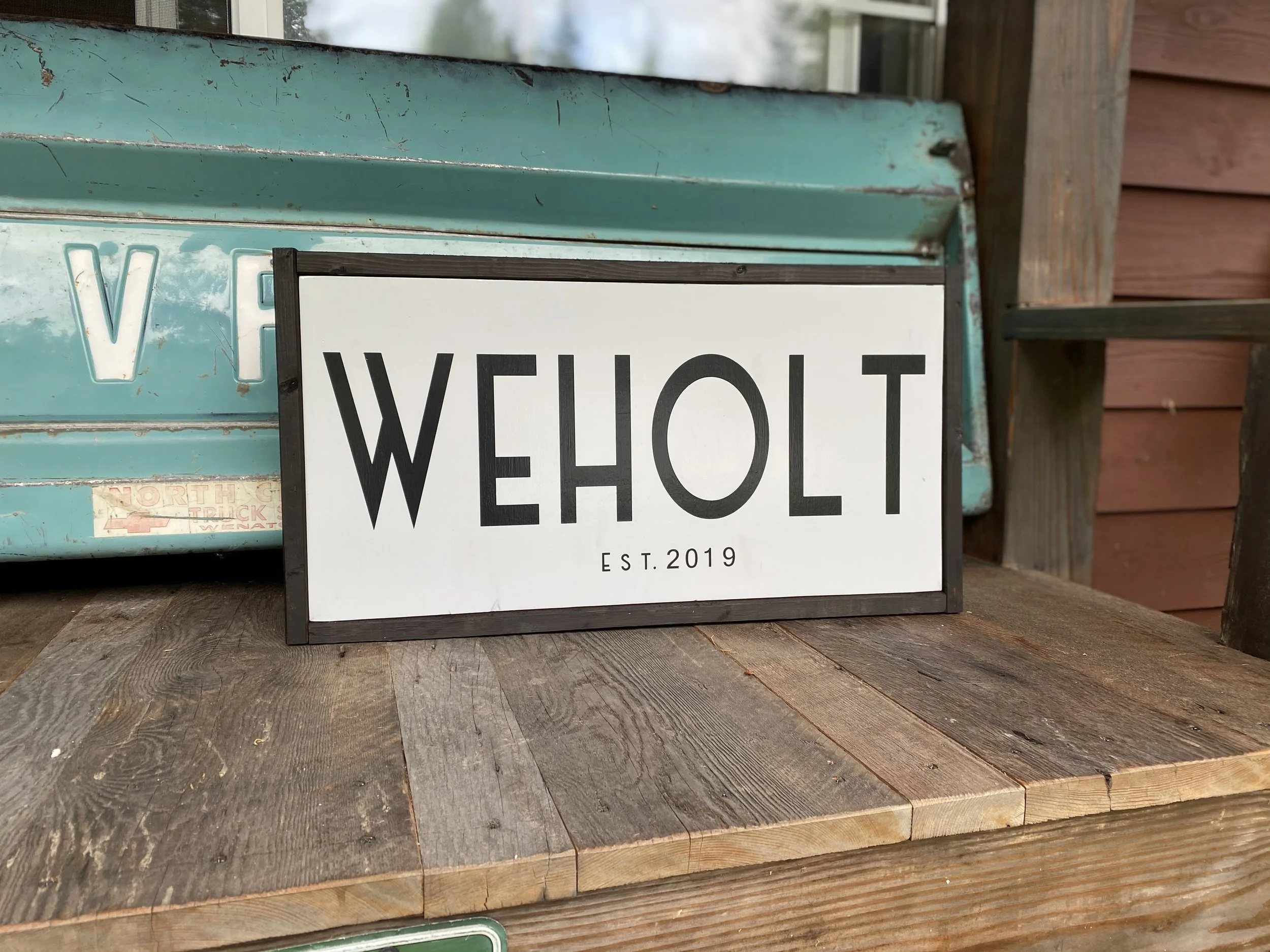 Sign with 'WEHOLT' in large black letters and 'EST. 2019' underneath, placed on a wooden surface against an old turquoise metal sign and a wooden wall.