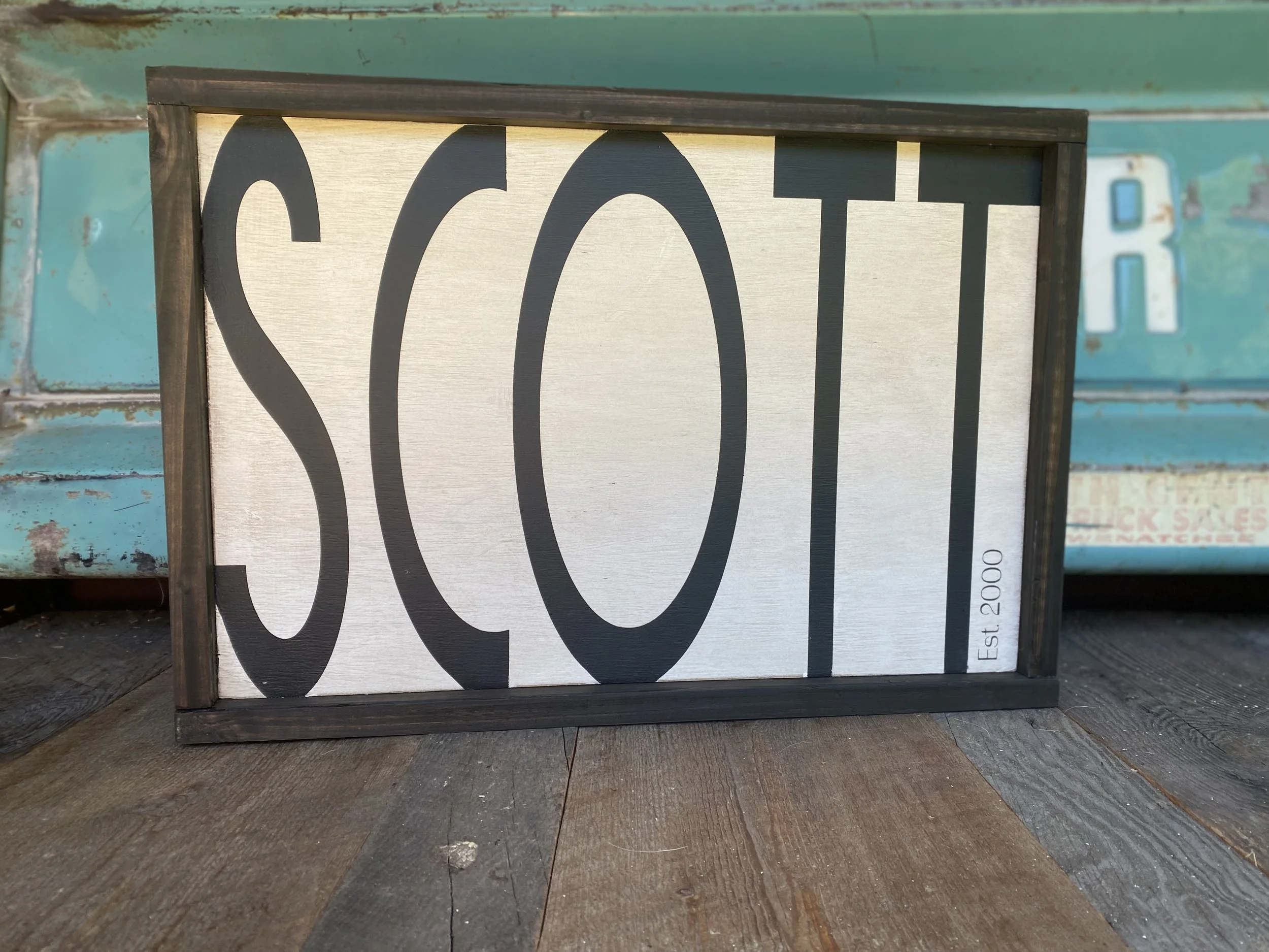 A rectangular sign with a black frame displaying the word 'SCOOT' in large black letters on a white background, with 'Est. 2000' written vertically in small letters.
