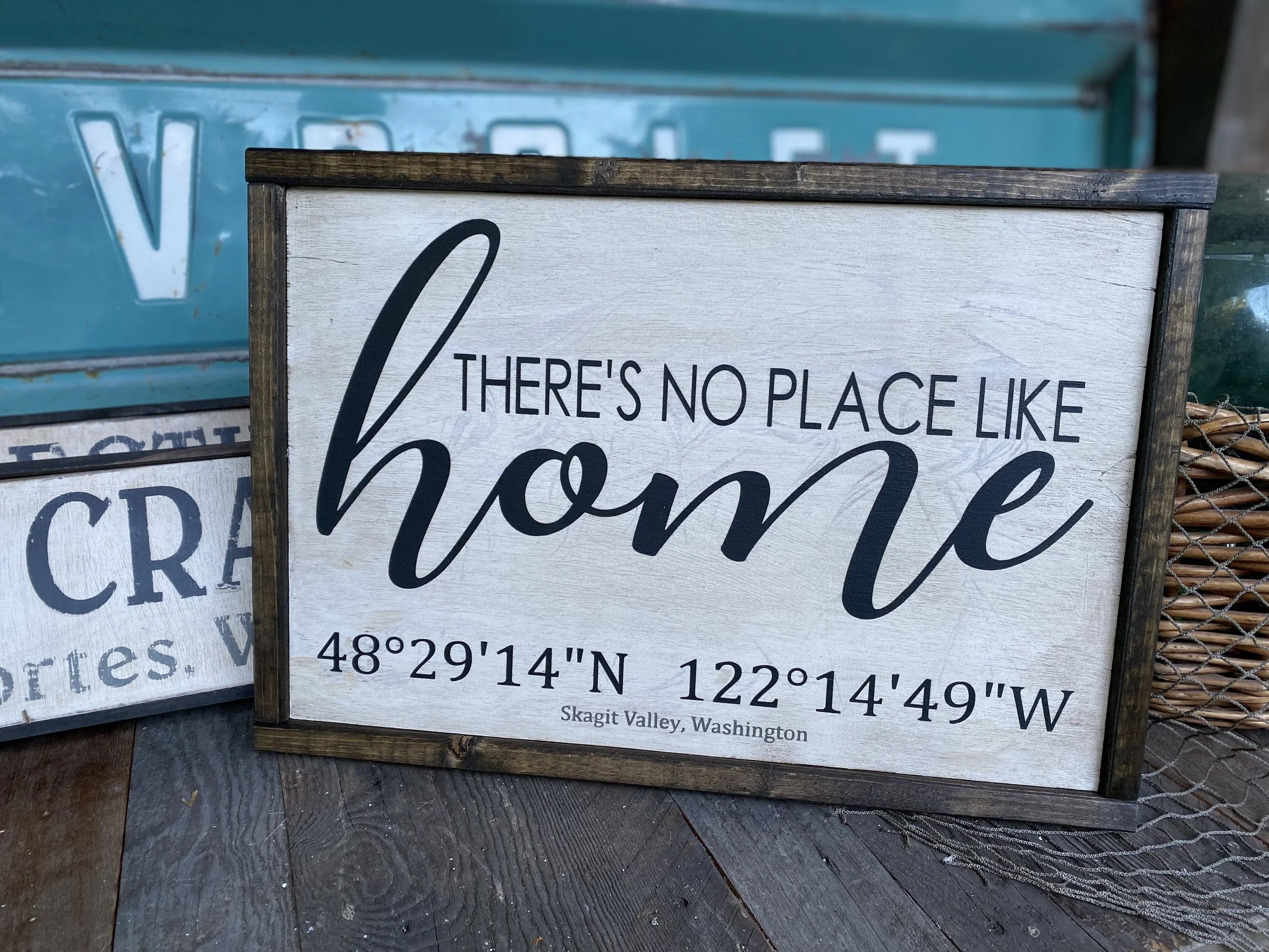 A wooden framed sign that reads, 'There's no place like home,' with coordinates 48°29'14"N 122°14'49"W and 'Skagit Valley, Washington' at the bottom. The background includes a teal-colored vintage truck and other rustic decor.