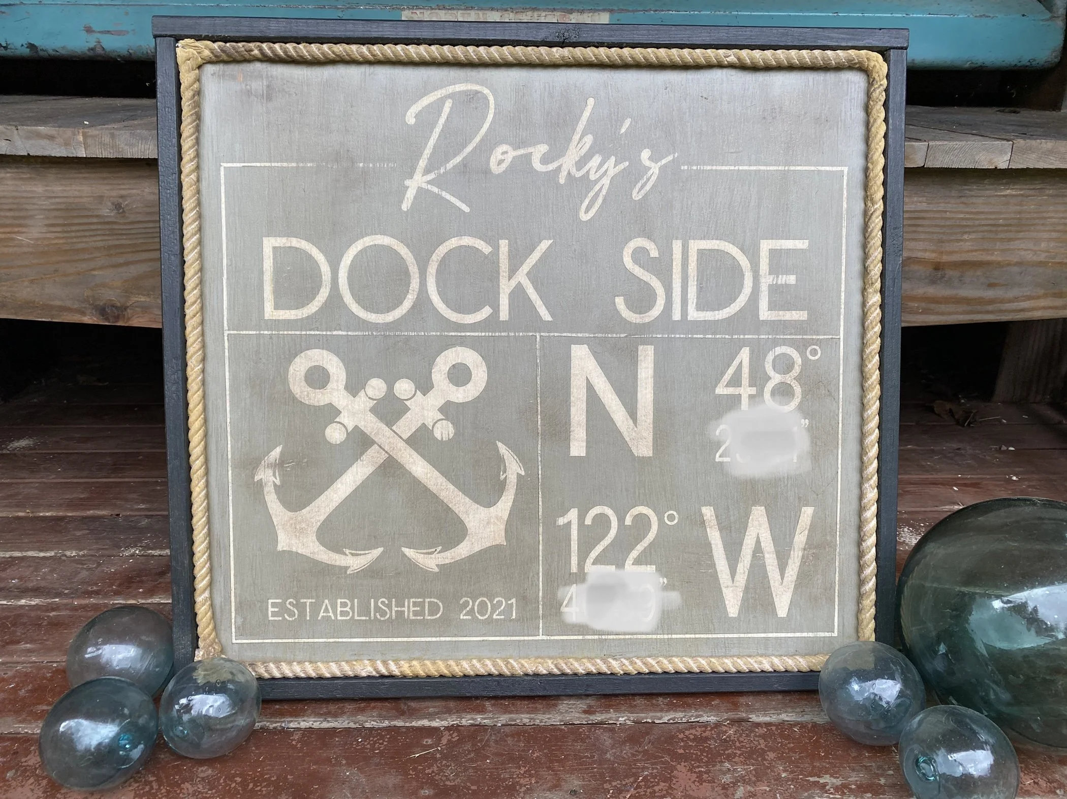A decorative sign with nautical theme, reading 'Rocky's Dock Side' with crossed anchors and established date 2021. It displays GPS coordinates, 48 degrees North and 122 degrees West, with a frame of thick rope and surrounded by glass decorative orbs.