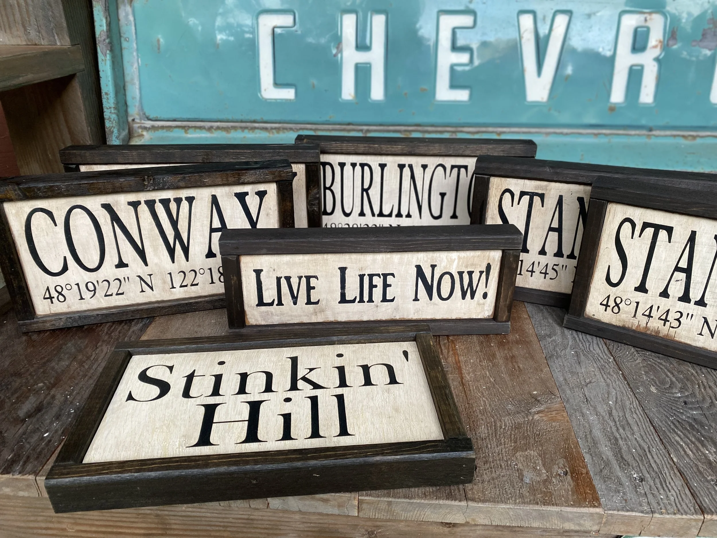 Collection of decorative rustic signs with city names, coordinates, and motivational phrases, displayed on wooden surfaces, with a faded turquoise Chevrolet sign in the background.