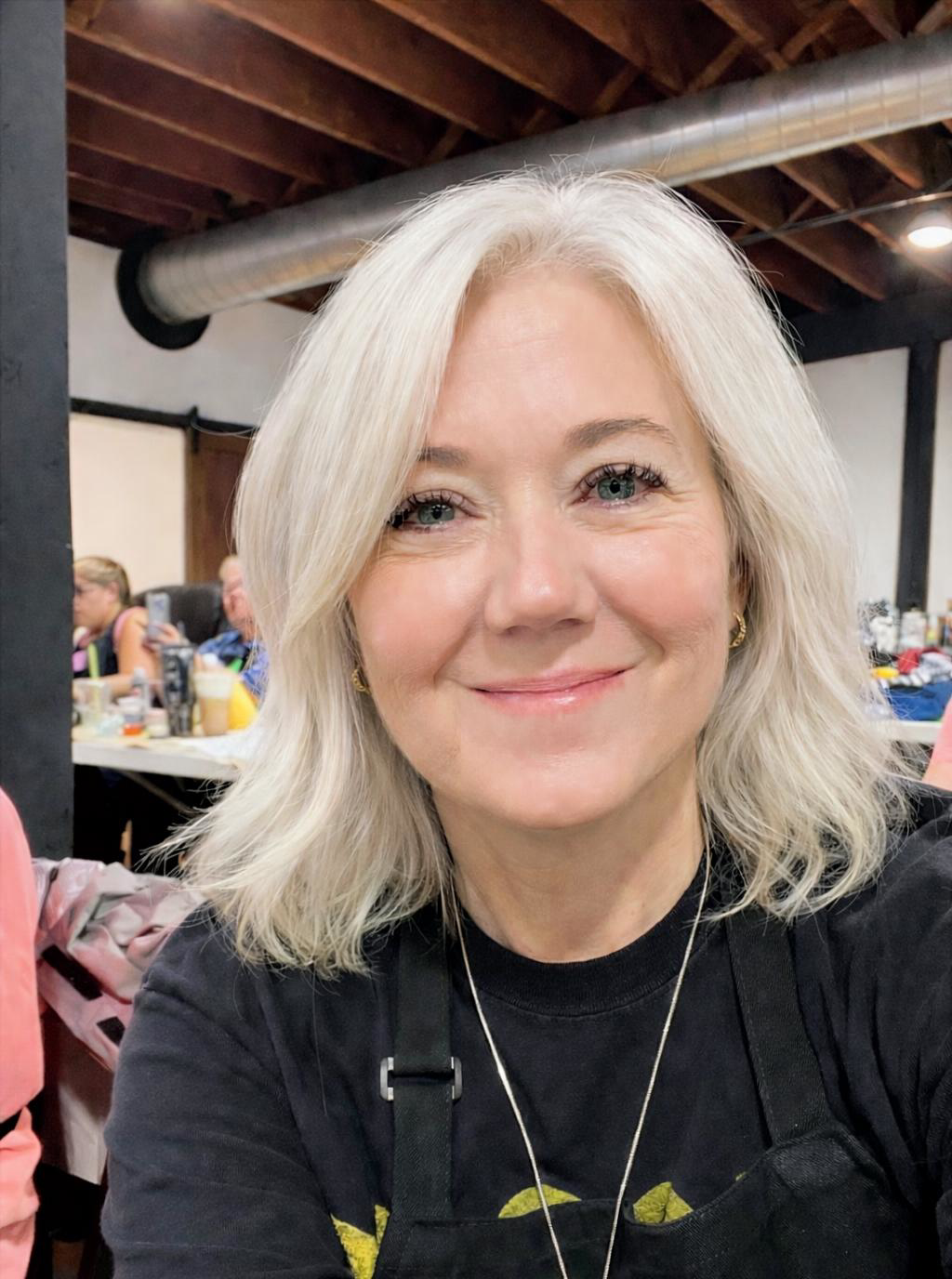 A smiling woman with blonde, shoulder-length hair and blue eyes, wearing a black top and a necklace, sitting in a room with a wooden ceiling and a visible metal vent pipe, with a background of a room with people and tables.