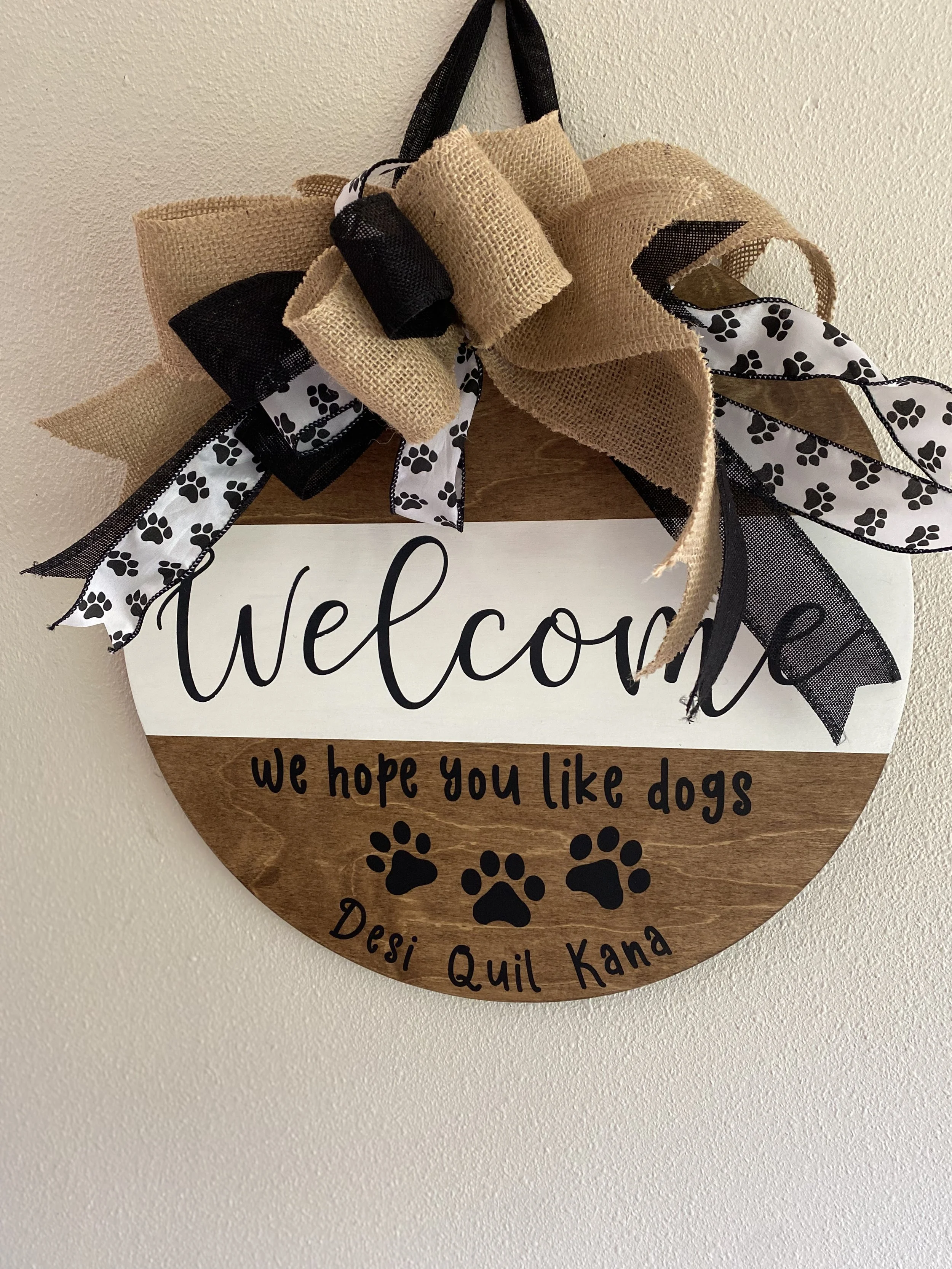 A round decorative sign hanging on a wall with a burlap and black ribbon bow on top. The sign reads 'Welcome' in large cursive letters, with the message 'We hope you like dogs' below, along with three black paw prints and the name 'Desi Quill Kana' a
