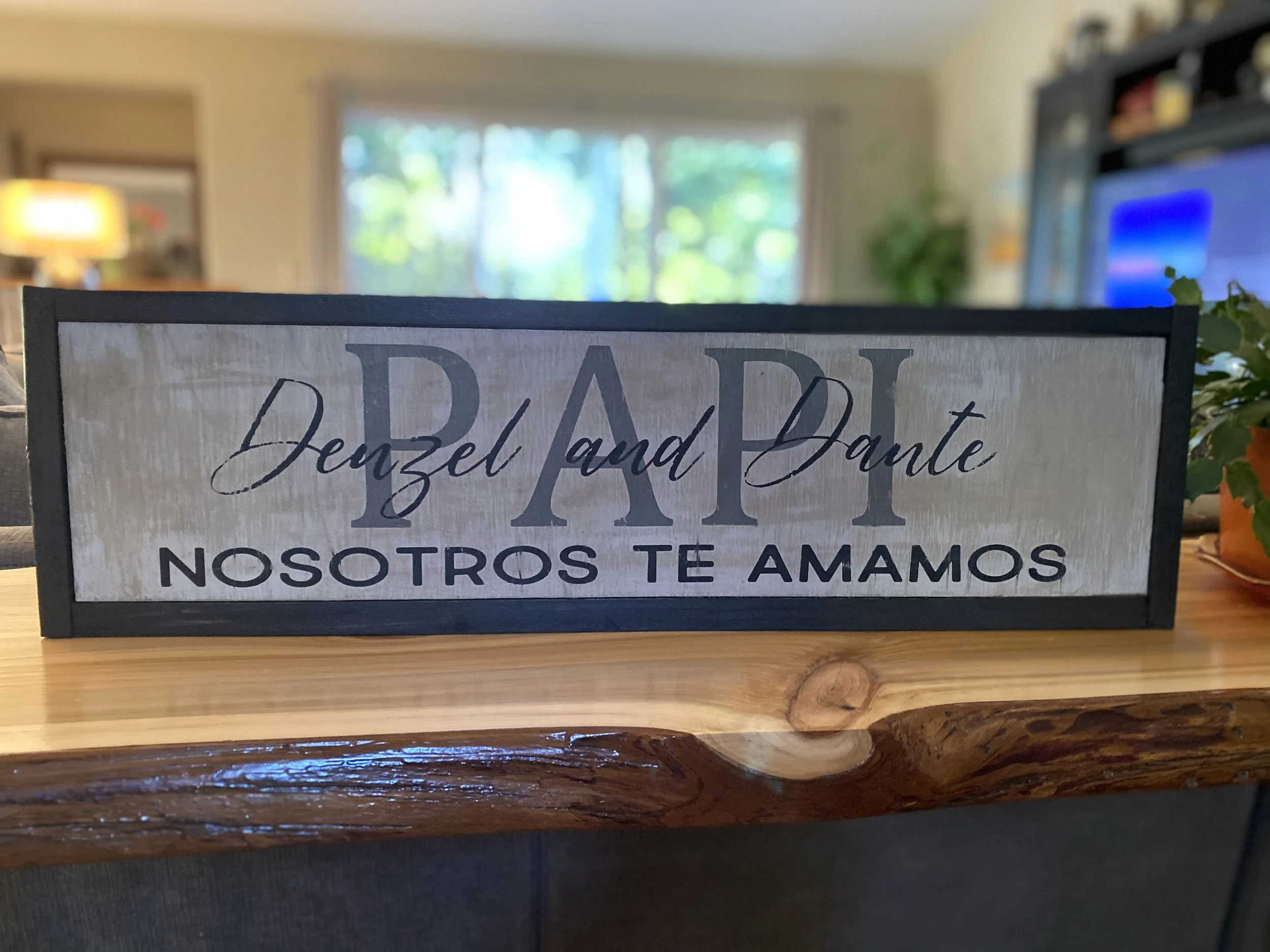 A decorative sign on a wooden table with the text "PAPA Denzel and Dante Nosotros Te Amamos," in a room with a window and TV in the background.