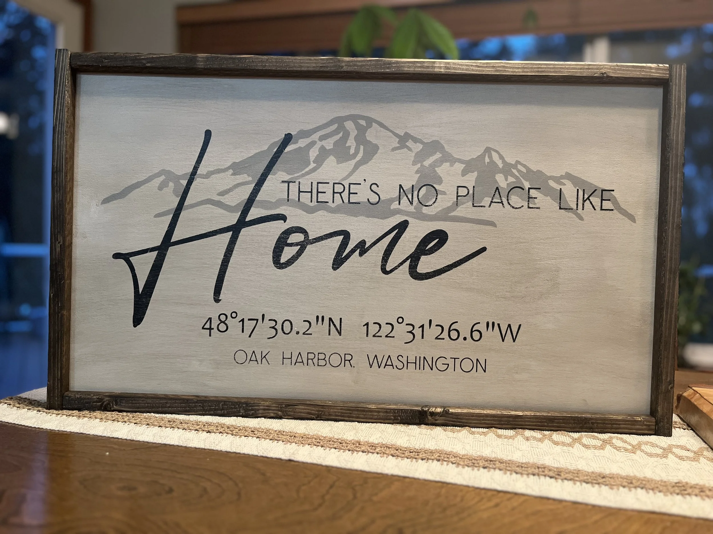Decorative wooden sign with image of a mountain range and text reading "There's no place like home", including geographical coordinates and location details for Oak Harbor, Washington.