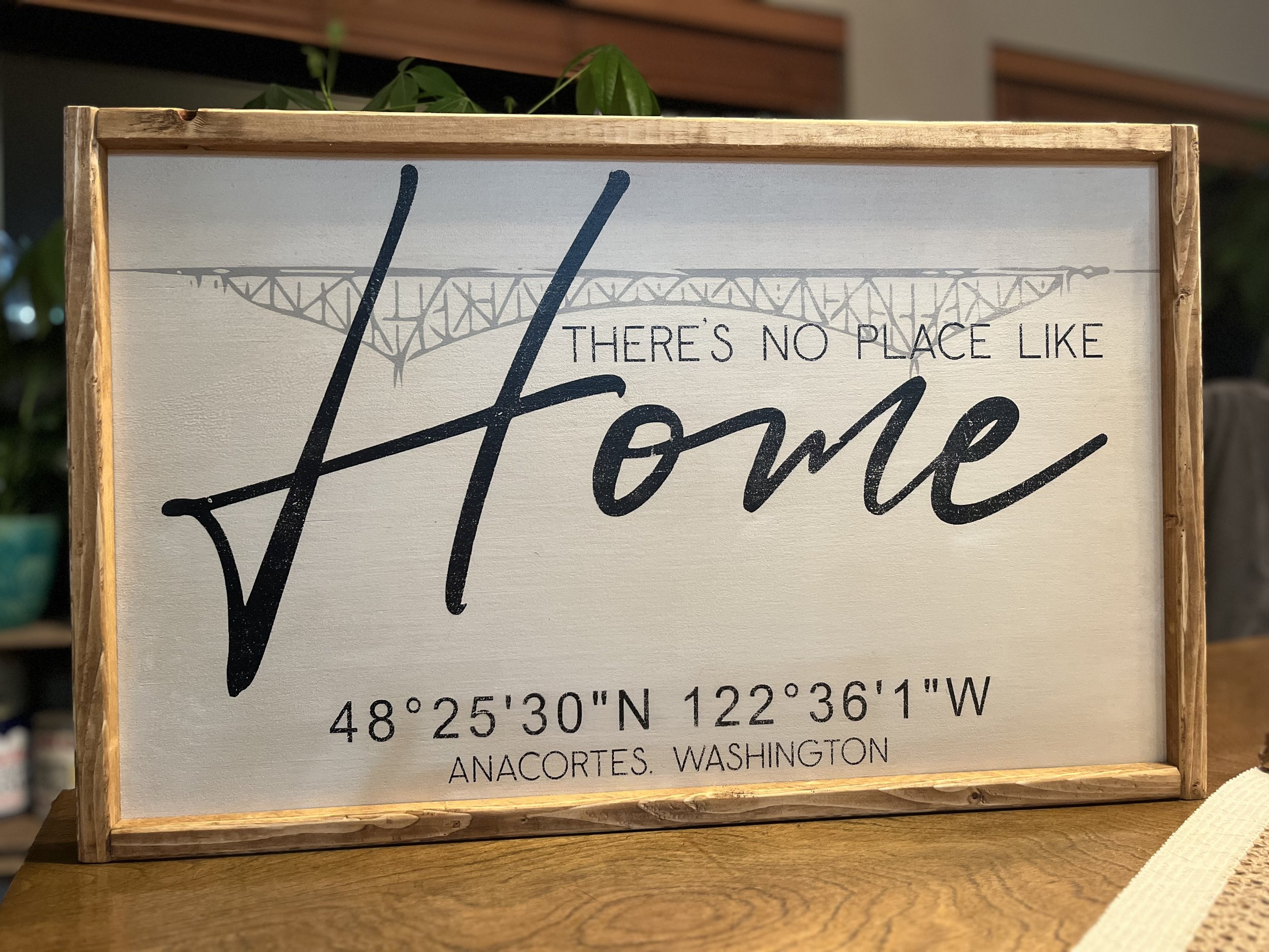 A decorative sign with the phrase "There's no place like home" featuring a background outline of a bridge, animated with coordinates "48°25'30"N 122°36'1"W" and location "Anacortes, Washington," framed with wood and placed on a wooden surface.