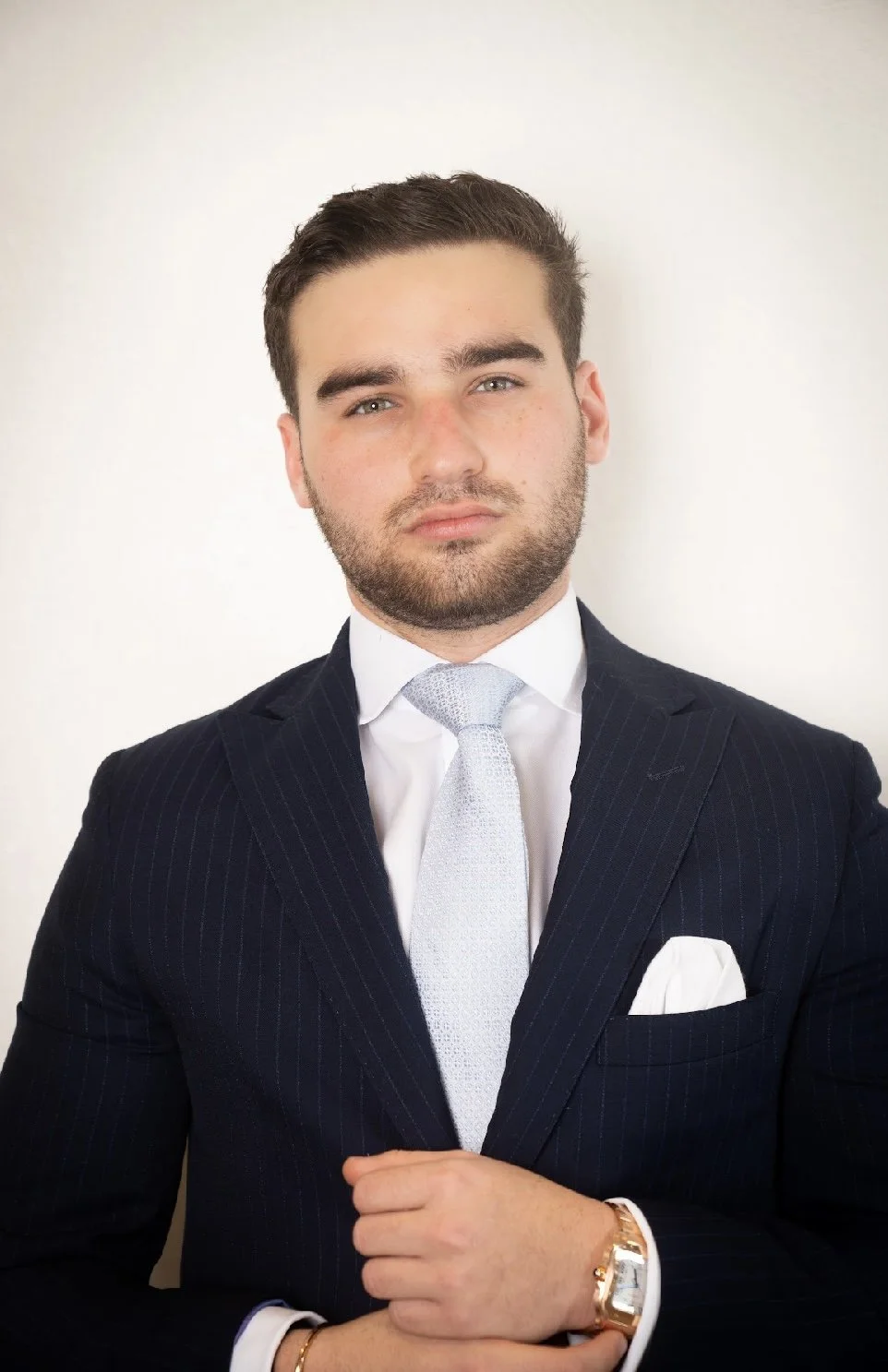 A young man in a dark pinstripe suit and silver tie, standing against a plain light-colored background, with a well-groomed appearance and a serious expression.