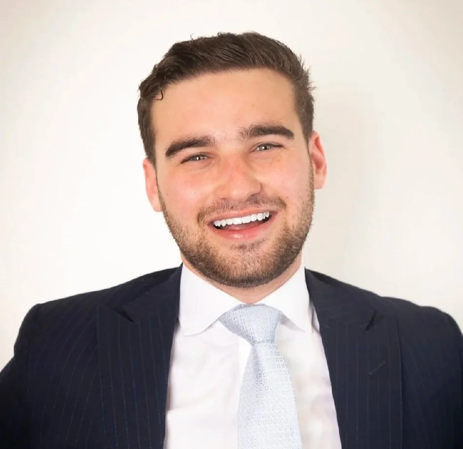 A young man with brown hair and a beard, smiling, wearing a dark suit jacket, white shirt, and a light-colored tie, standing against a plain light background.