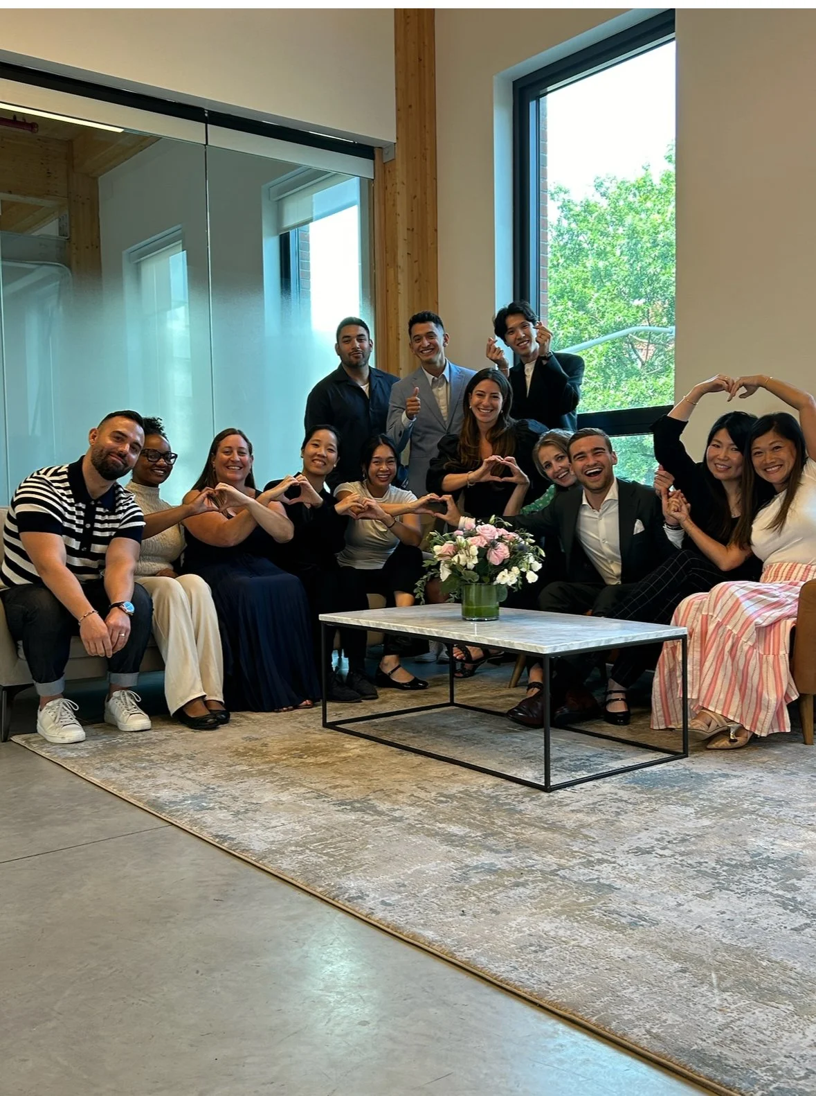 A group of people sitting and standing together in a room with large windows, making heart shapes with their hands and smiling.