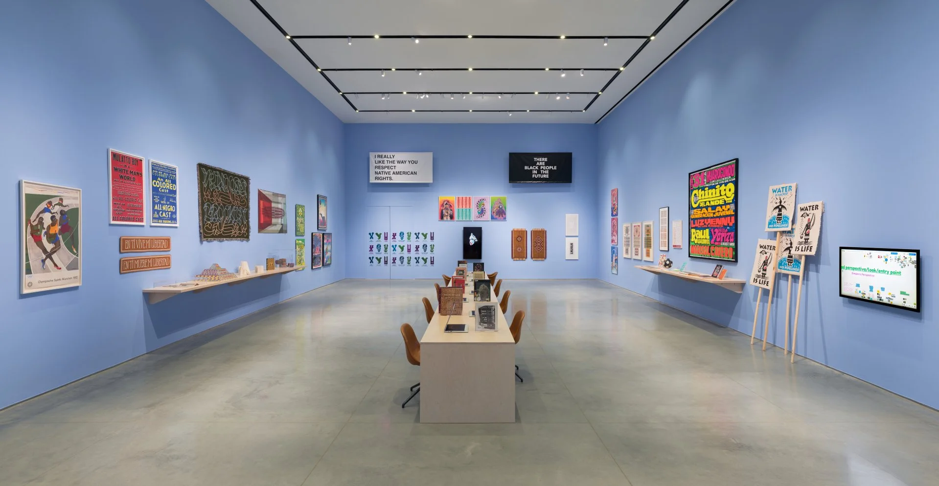 An art gallery with blue walls displaying various colorful artworks, posters, and framed pieces. There are two digital screens, one featuring text and the other showing abstract graphics. A central table with chairs holds books and displays, and a shelf along the left wall contains small sculptures and items.