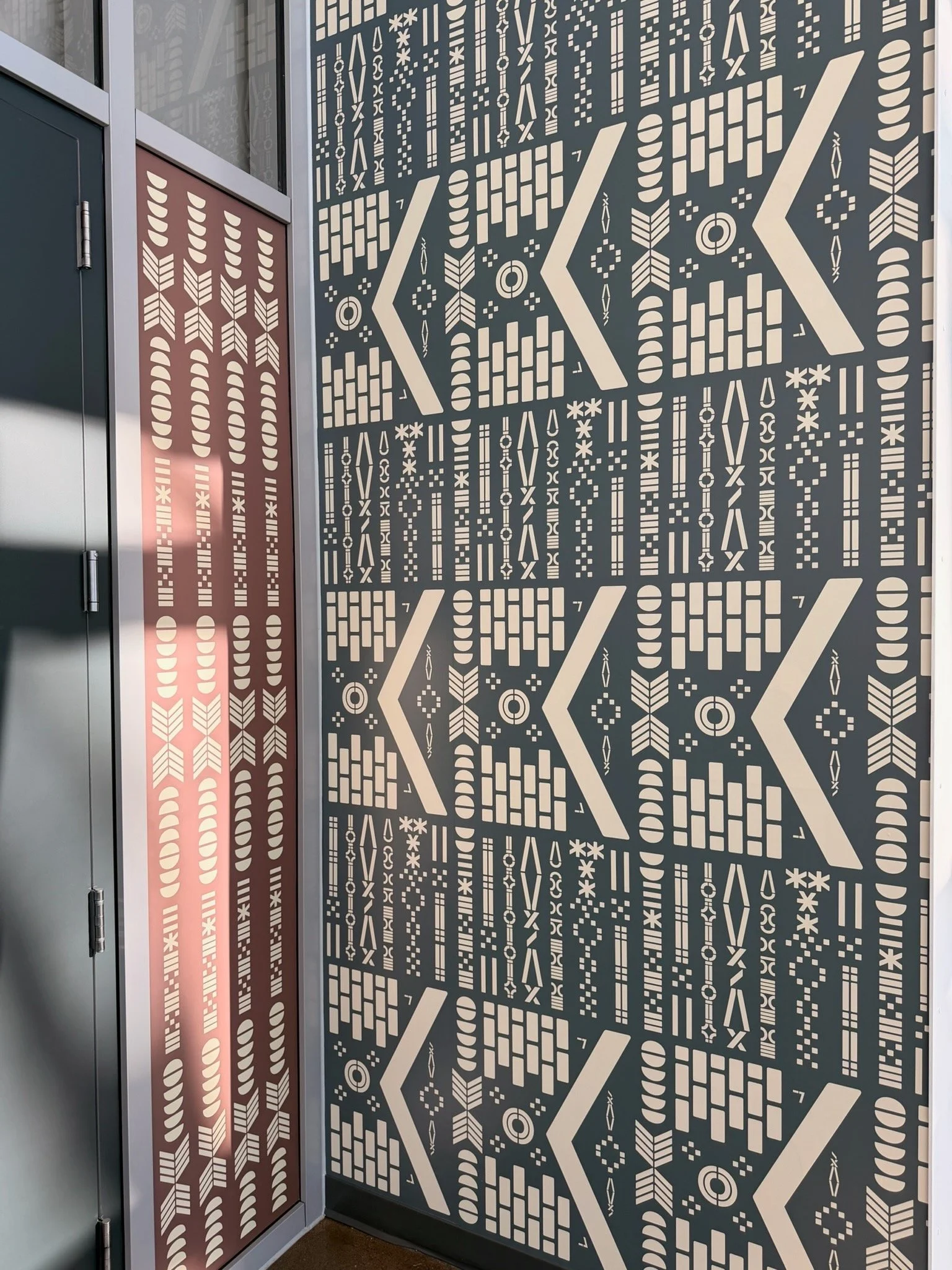 Decorative wall with geometric patterns and a partly visible door with similar design, featuring arrows, lines, and shapes in beige on a dark background.