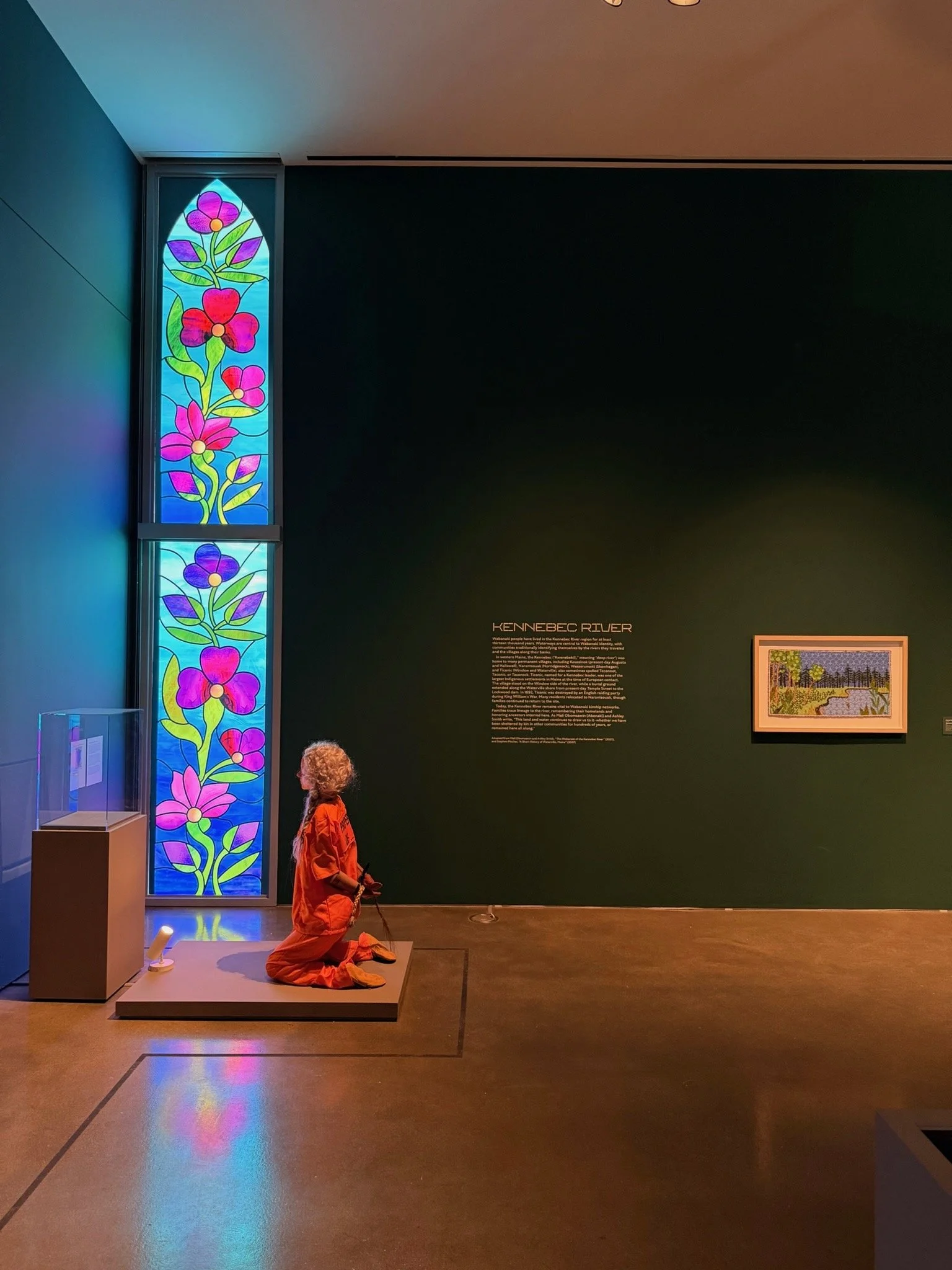 A museum exhibit featuring a colorful stained glass window with pink and purple flowers and green leaves, a sitting mannequin dressed in an orange outfit, and a dark green wall with text about the Kennebec River and a framed landscape painting.