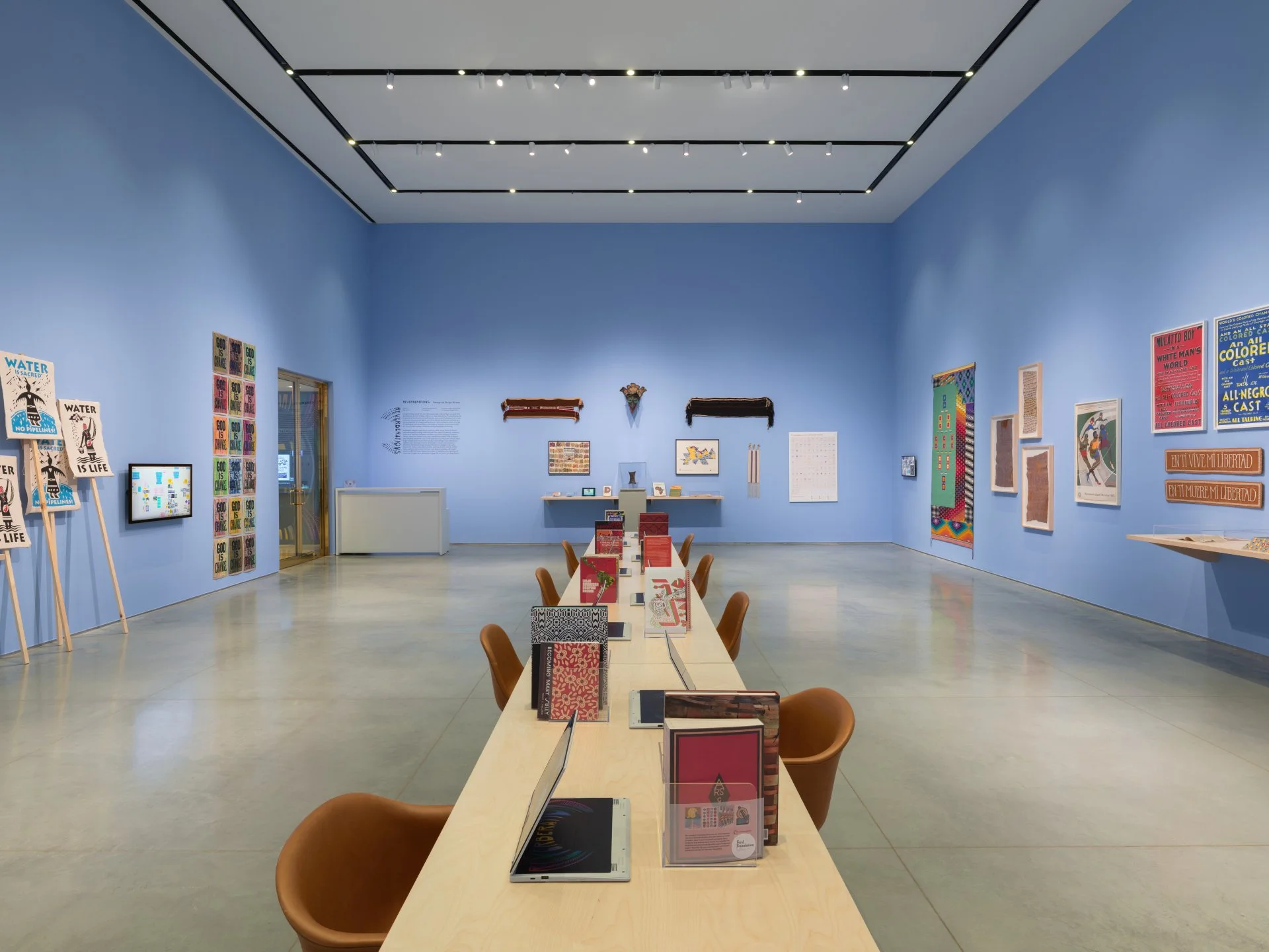 Art gallery with blue walls, colorful artwork on display, a long table with books and tablets, and brown chairs.