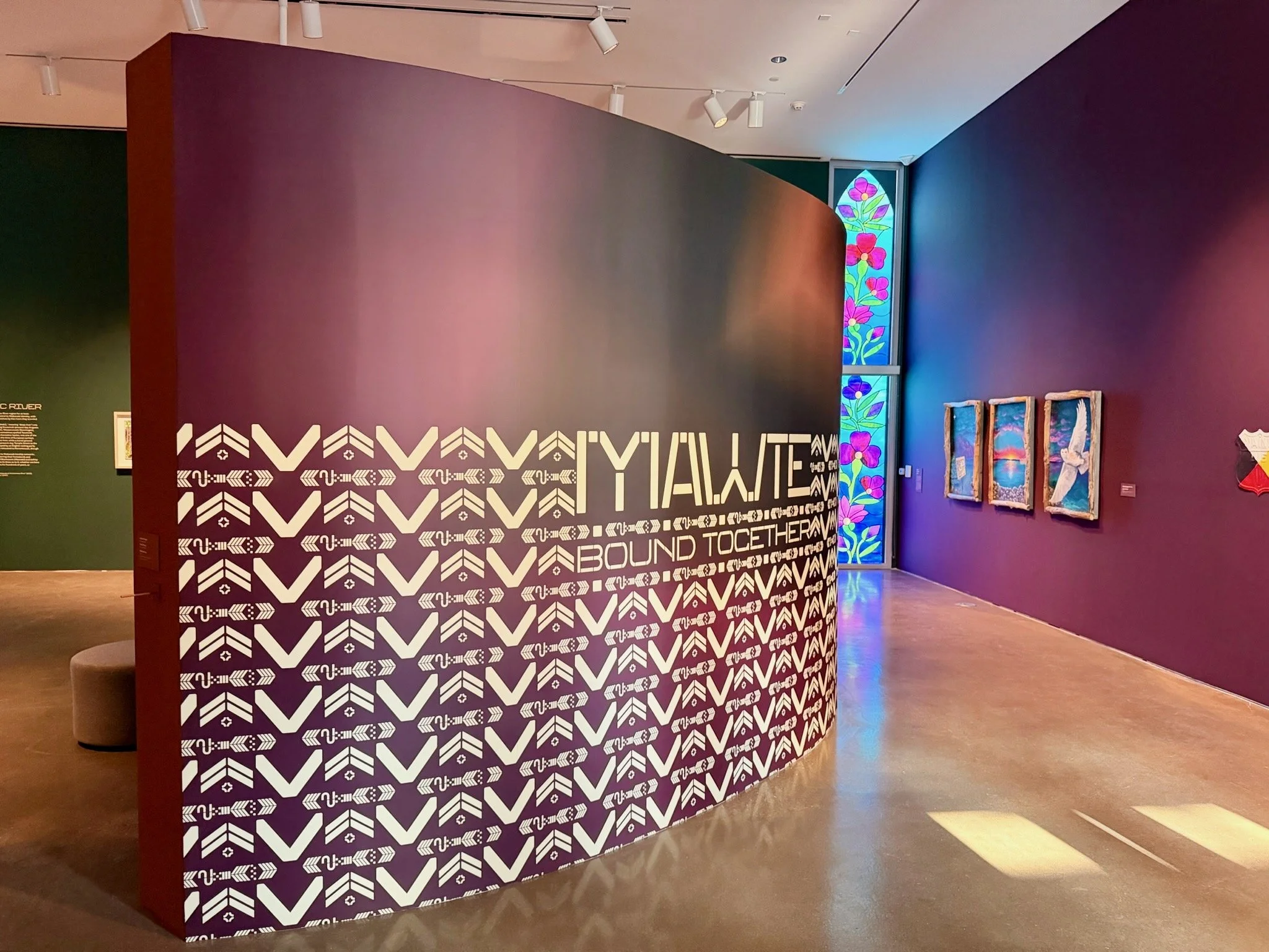 Art exhibit in a gallery with a colorful stained glass window, three paintings on a purple wall, and a decorated partition with text and geometric patterns.