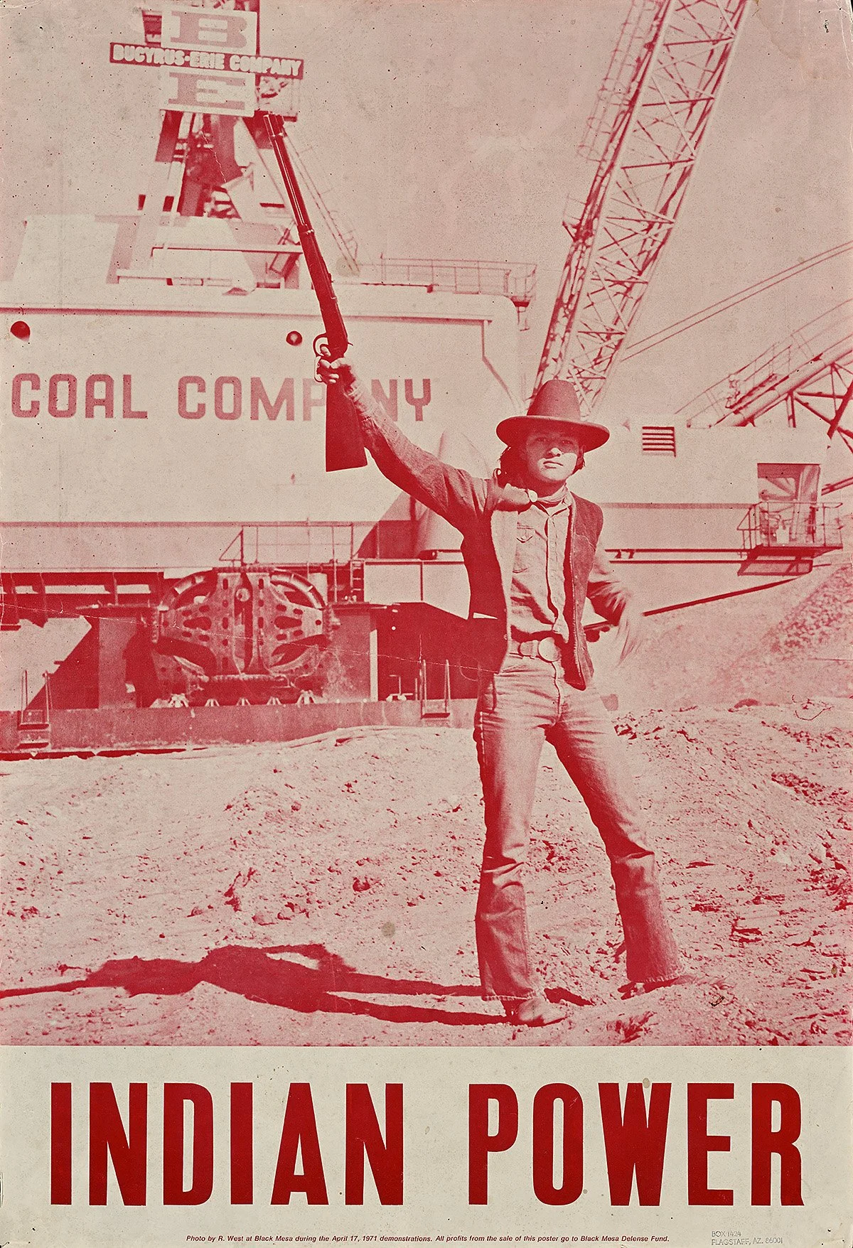 A person dressed like a cowboy, wearing a hat, sunglasses, and western clothes, standing in a dirt area at a political protest with a raised rifle. In the background, there is a large industrial mining or construction site with a crane and a sign that reads 'Coal Company', and a banner above displaying numbers and text.