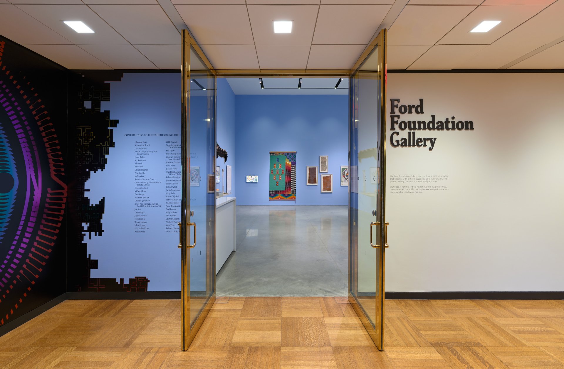 Entrance to the Ford Foundation Gallery showing an interior with artworks on blue walls, glass double doors, and a sign with the gallery's name.