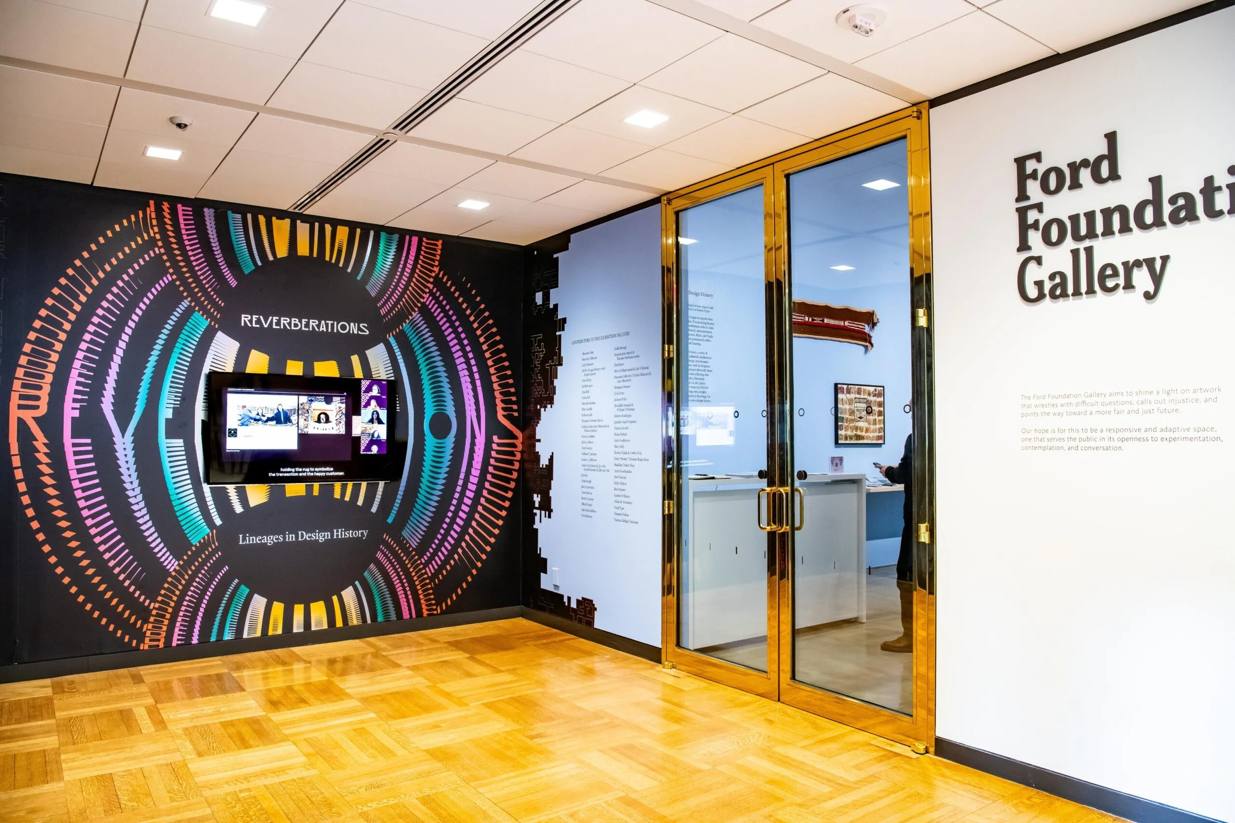 Entrance to the Ford Foundation Gallery with a colorful circular design on the wall on the left side, a digital display screen, and a white wall with the gallery name in large black letters on the right, inside a modern indoor space with wooden flooring.