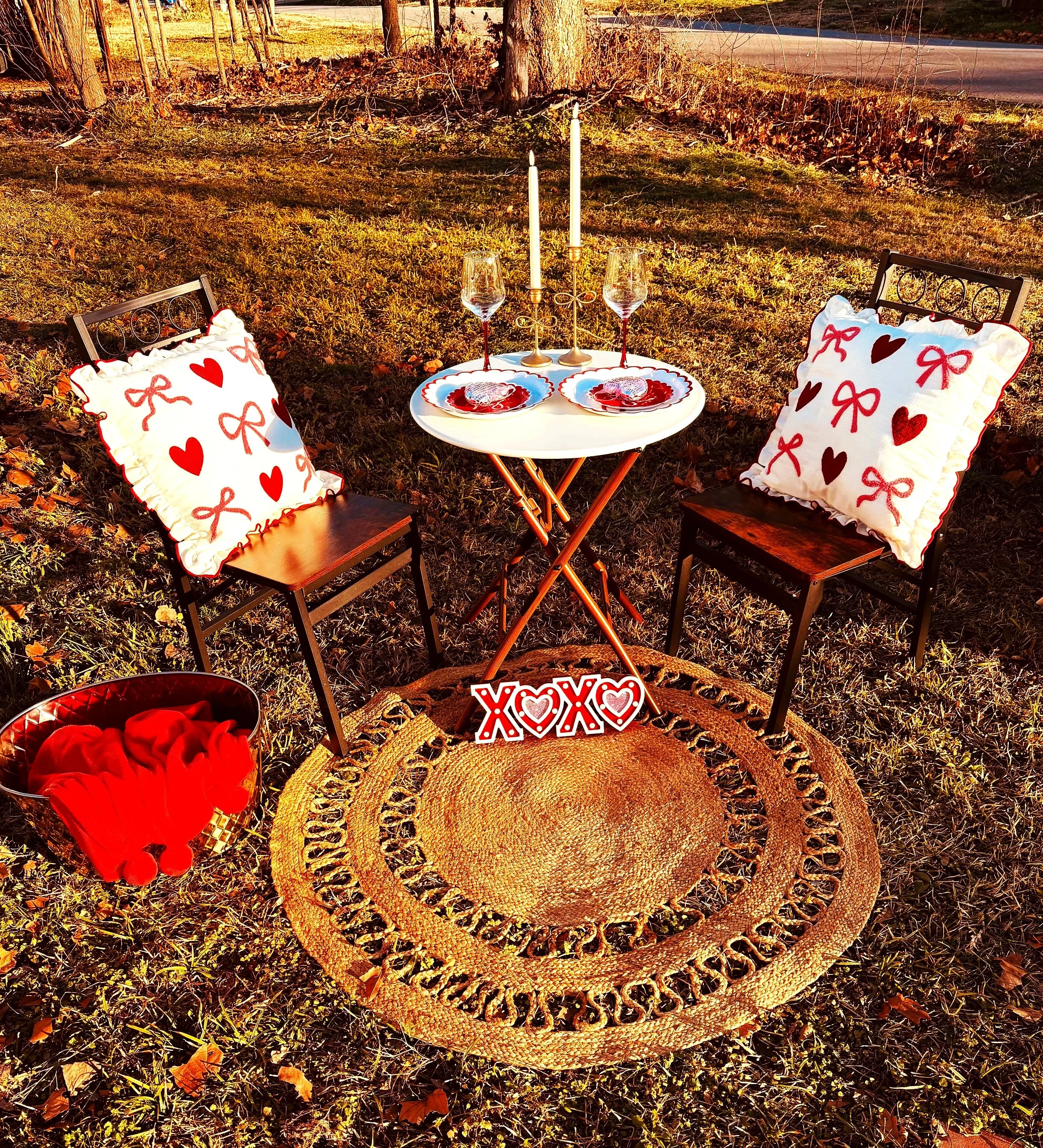 A romantic outdoor setup with two chairs, each with a pillow decorated with red hearts and bows. A small round table is set with two wine glasses, plates, and candles, decorated with a 'XOXO' sign on a woven rug on the ground, in a grassy area near a road.
