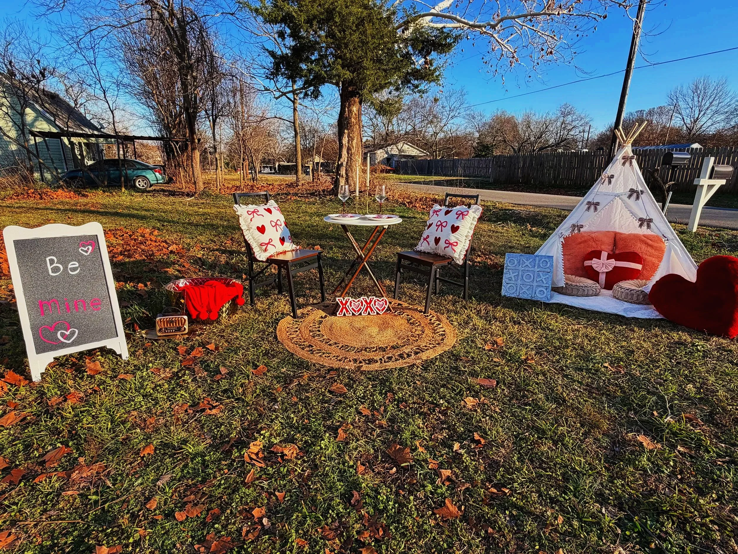 Decorative Valentine's Day setup in a backyard with a small white tent, heart-shaped pillows, a small round table with chairs, a sign that says 'Be mine,' and various Valentine's Day themed decorations.