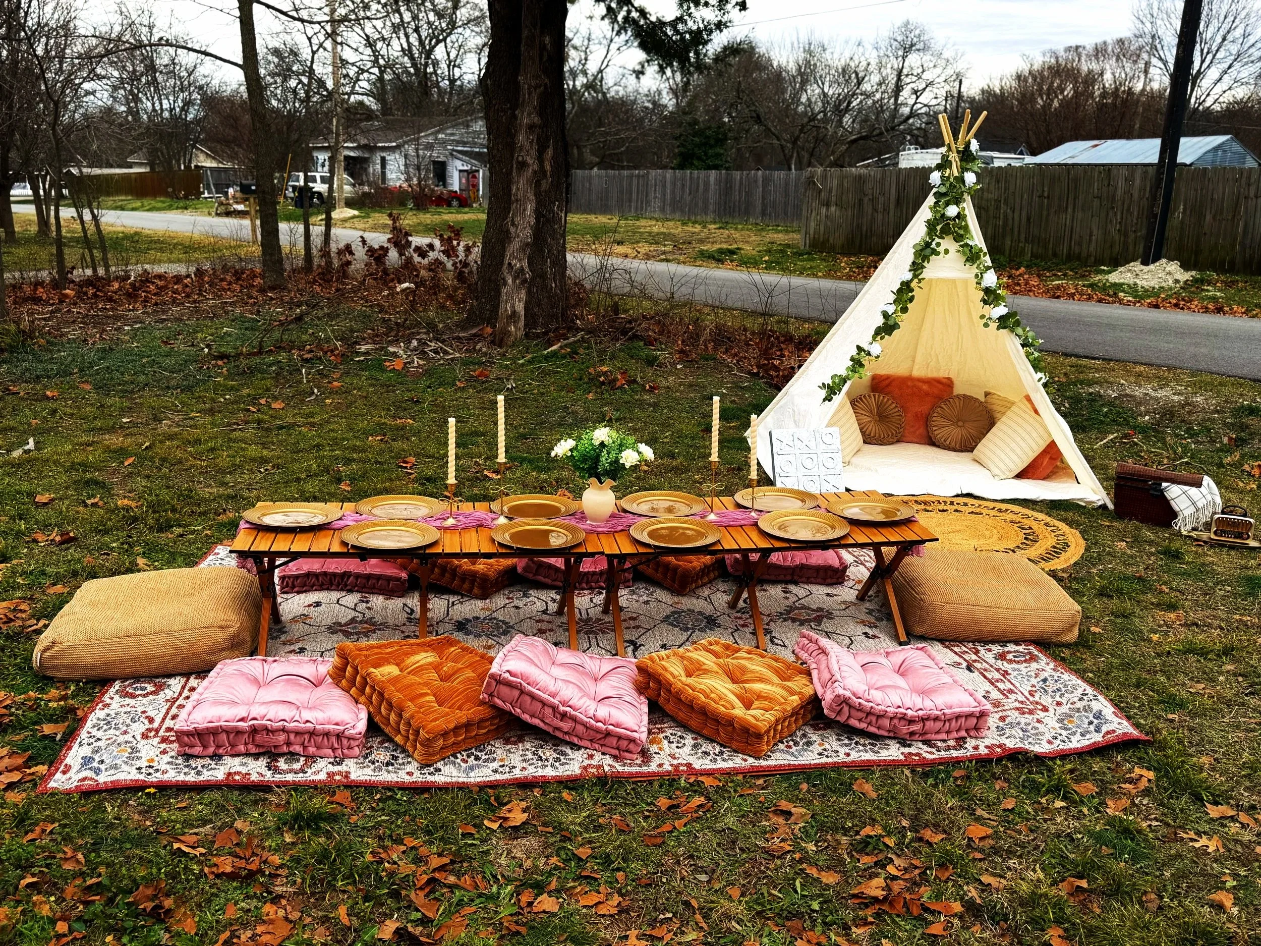 Outdoor bohemian-style picnic setup with a low table, cushions, pillows, and a white teepee decorated with greenery, set on a grassy yard surrounded by trees and a wooden fence.
