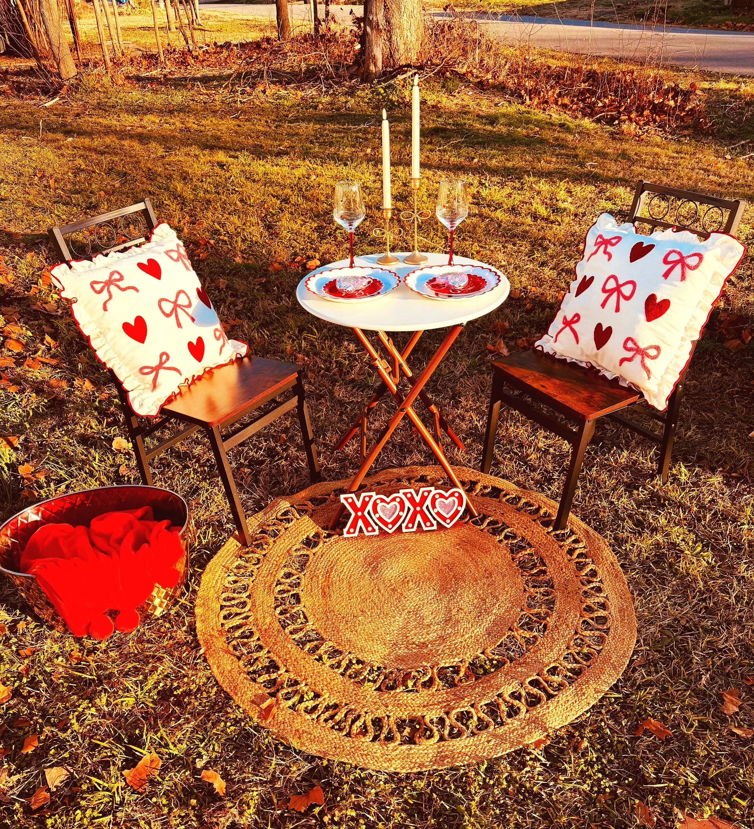 Outdoor romantic setting with two chairs, cushions with red hearts and bows, a small round table with candles, glasses, and heart-shaped plates, a round woven rug, a decorative "XOXO" sign, and a bucket of red cloth, all illuminated by warm sunlight.