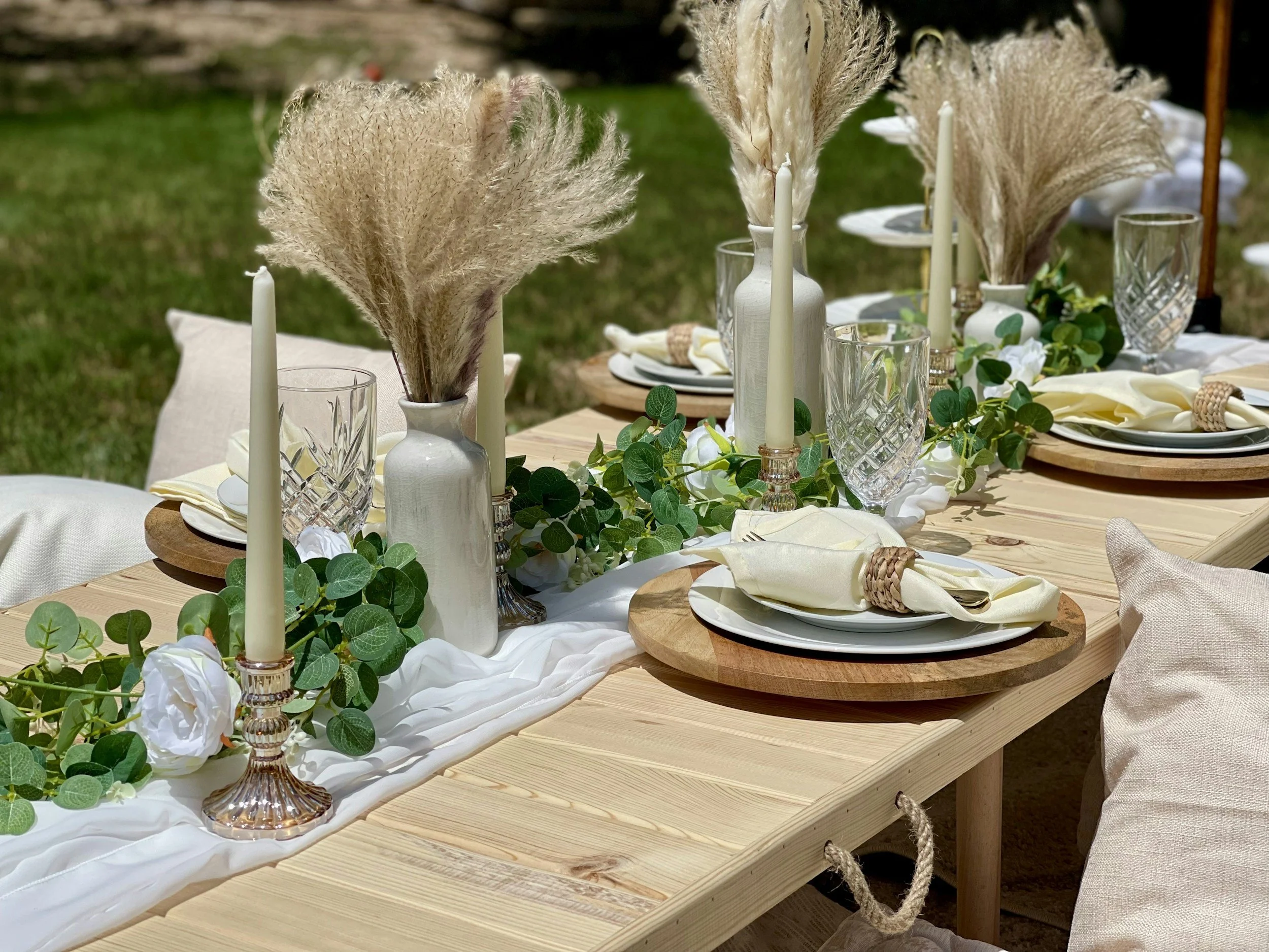 Decorative outdoor table setting with candles, pampas grass, and greenery on a wooden table with cream plates and napkins.