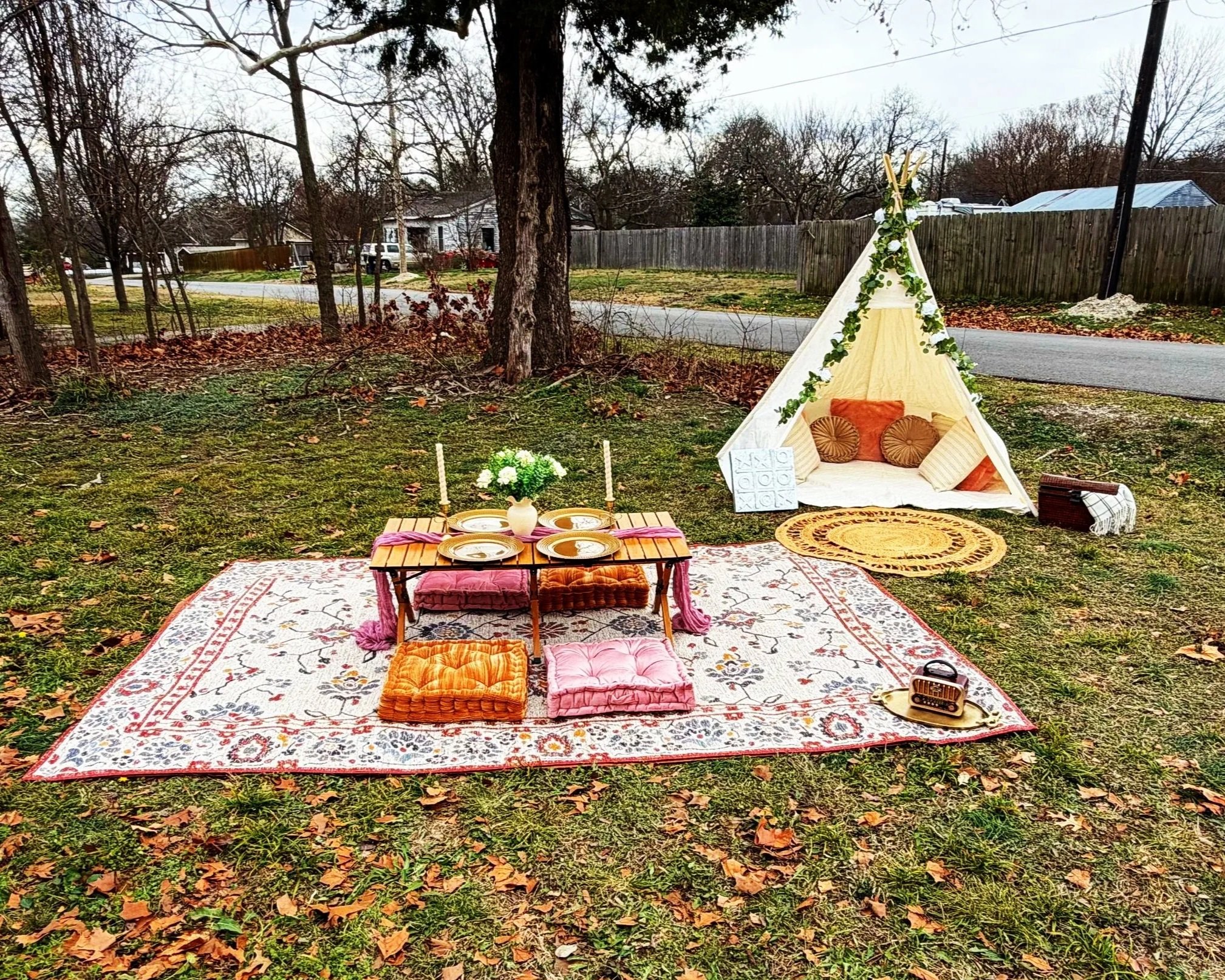 Outdoor bohemian-style picnic setup with a colorful rug, low table with plates, pillows, candles, and a teepee with cushions.