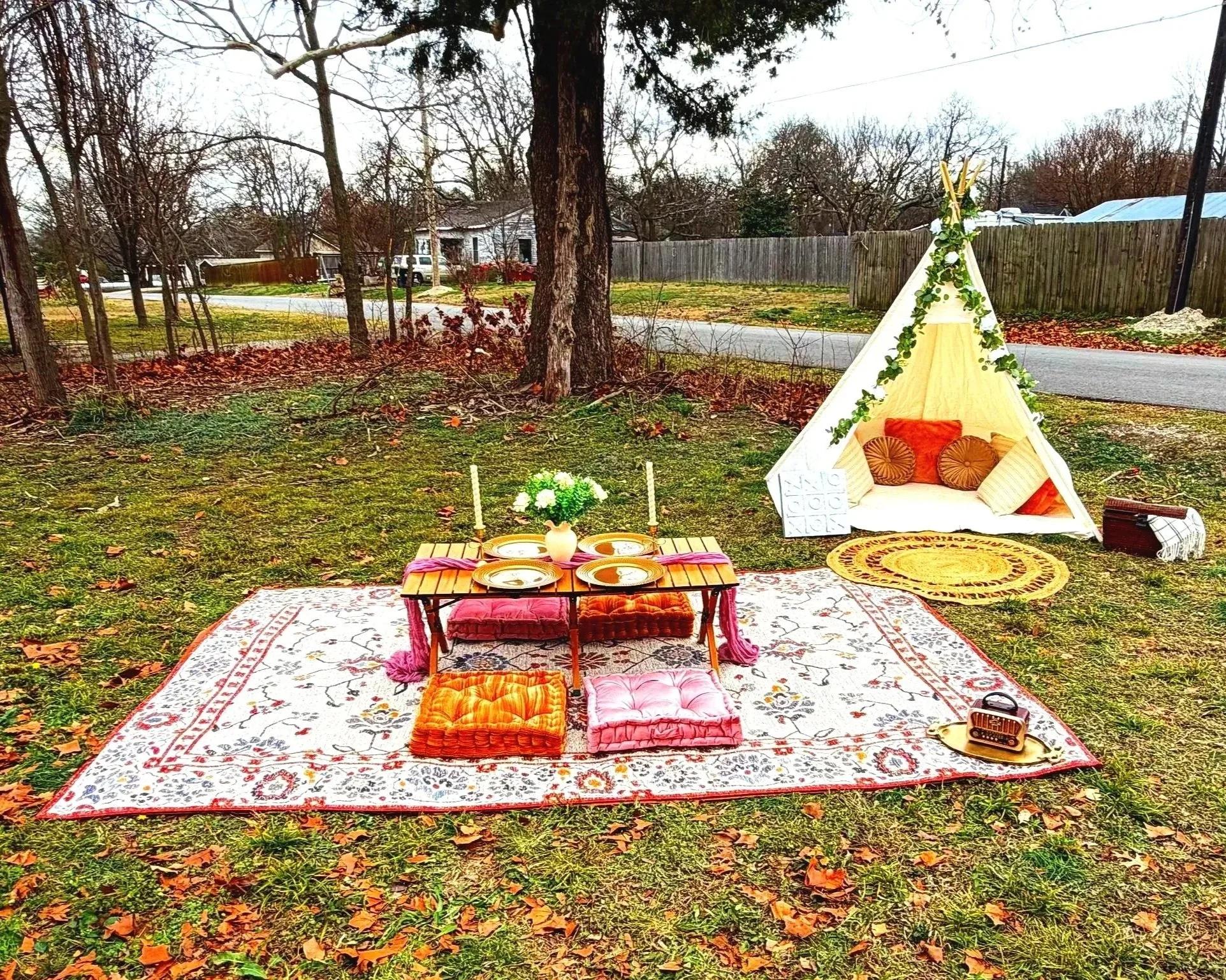 A cozy outdoor picnic setup on a vintage rug with cushions, a low wooden table, a flower vase, candles, and a teepee decorated with greenery, set in a backyard with trees and a fence.