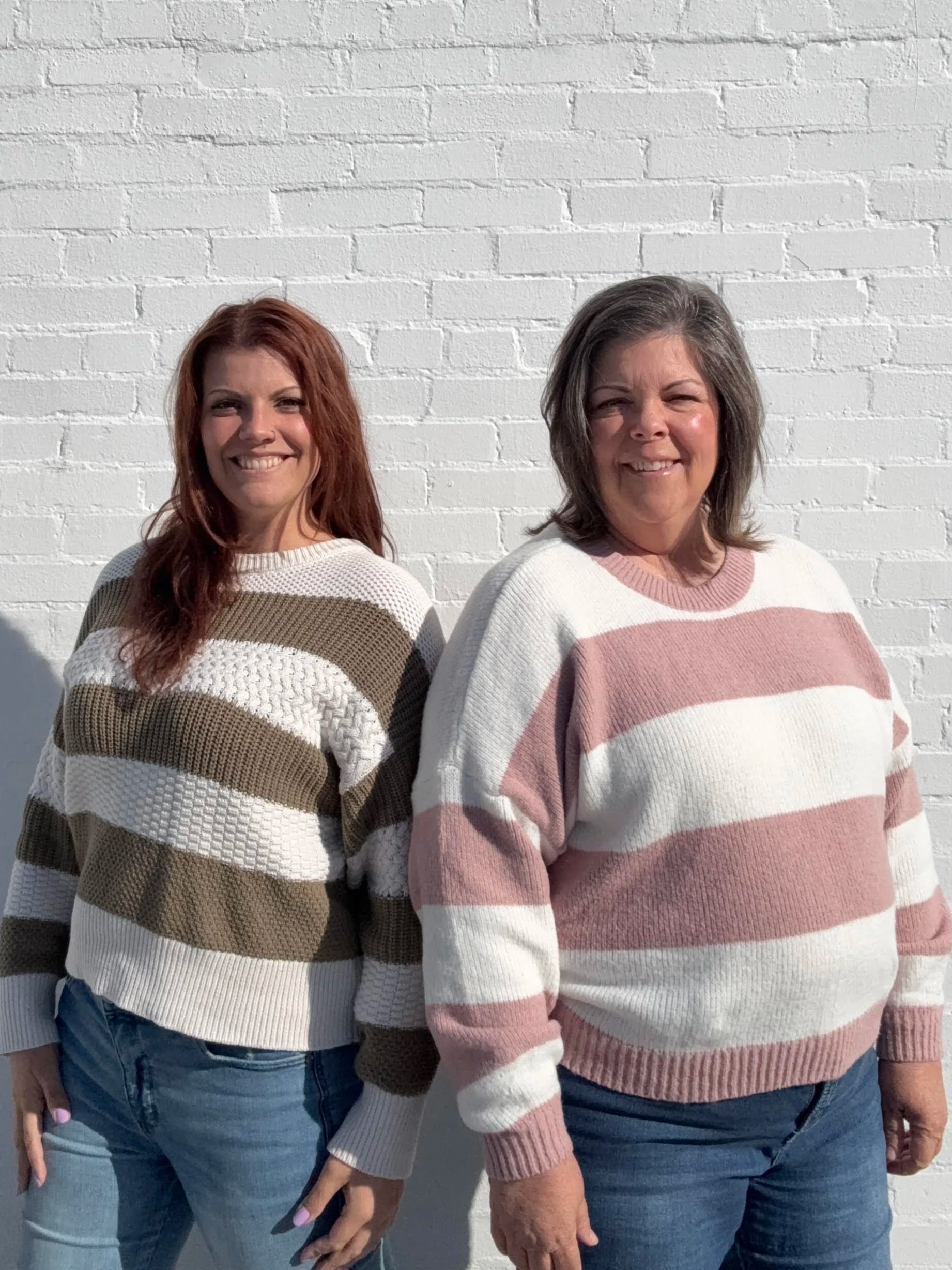 Two women standing side by side smiling against a white brick wall, wearing striped sweaters and jeans.