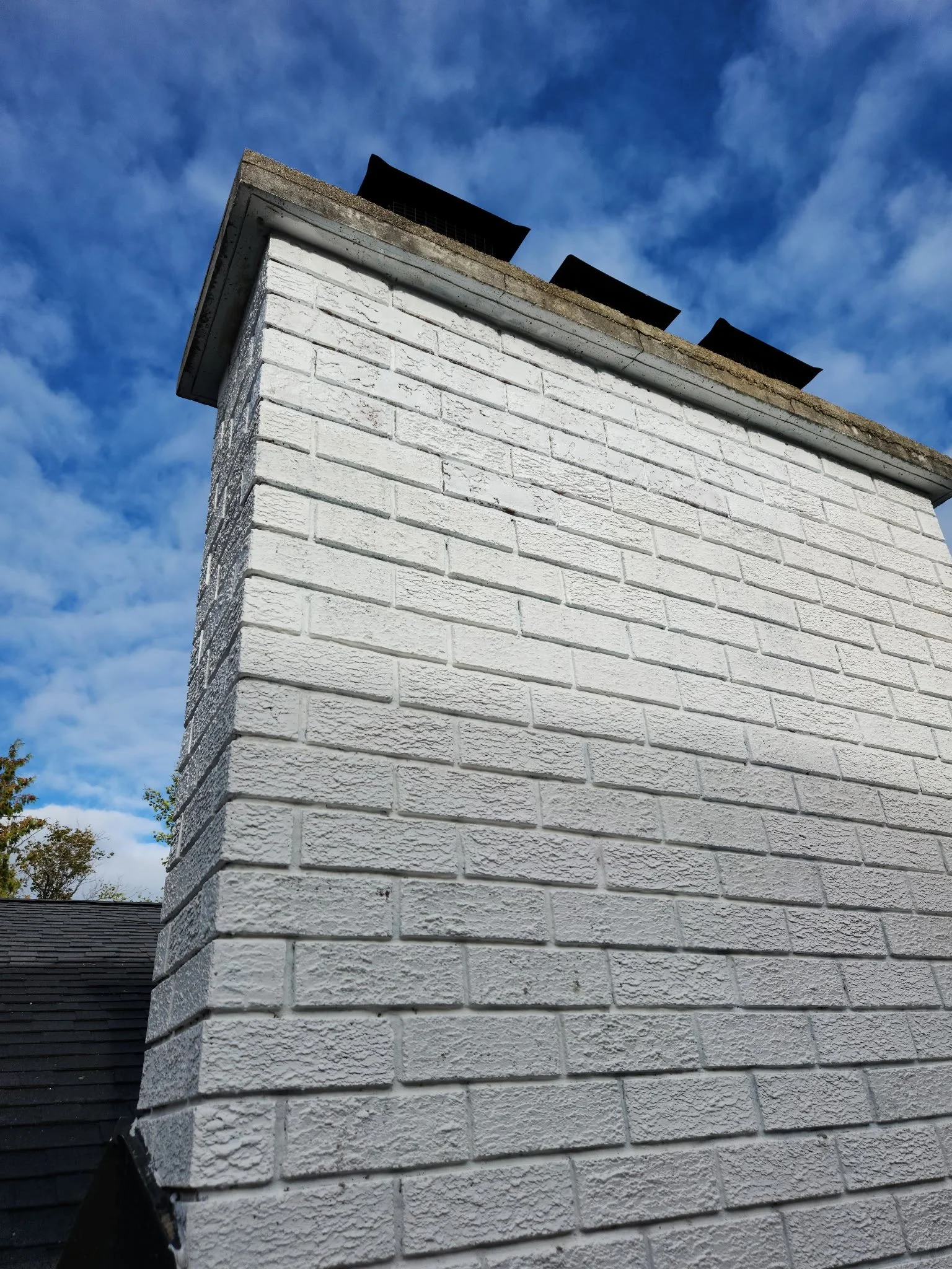 Close-up of a white brick chimney on a house with a dark roof, under a blue sky with scattered clouds.