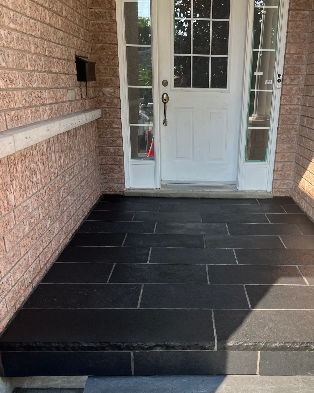 Front porch with black tiled flooring, white door with glass windowpanes, brick walls on each side, and a black mailbox mounted on the left wall.