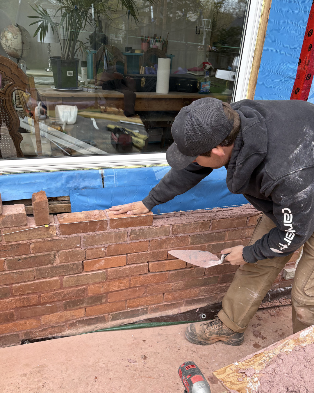 A man in a black hoodie and baseball cap laying bricks on a red brick wall outside a house, using a trowel.