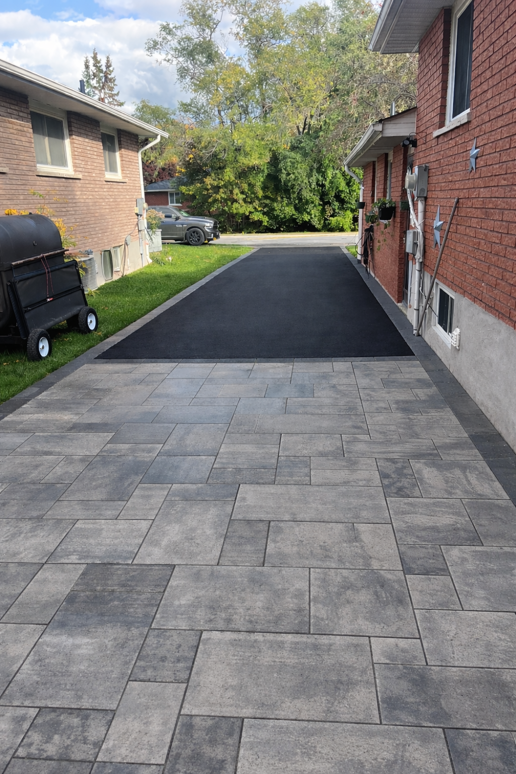 View of a driveway with a paved front area and a black asphalt extension, bordered by brick houses, with a black barbecue grill on the left and green grass on the right, trees in the background under partly cloudy sky.