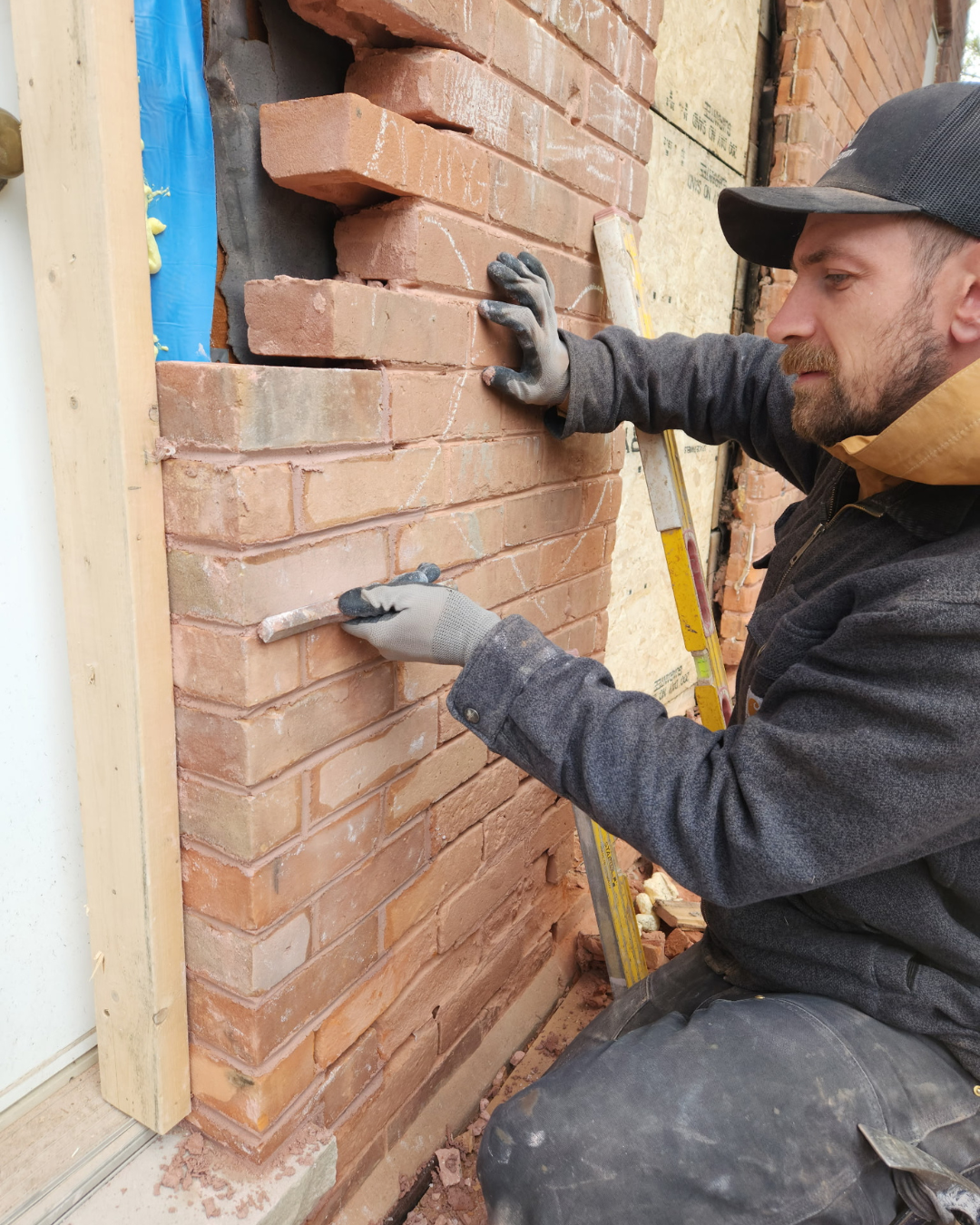 A man working on laying bricks on a wall, wearing gloves and dark clothing, with a yellow ladder beside him.