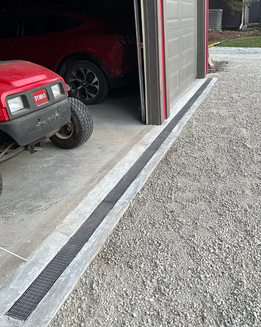 A garage with a red compact car parked inside, a black electric vehicle, and a red and black golf cart in front. The garage has a grated drain along the concrete floor edge, and gravel outside.