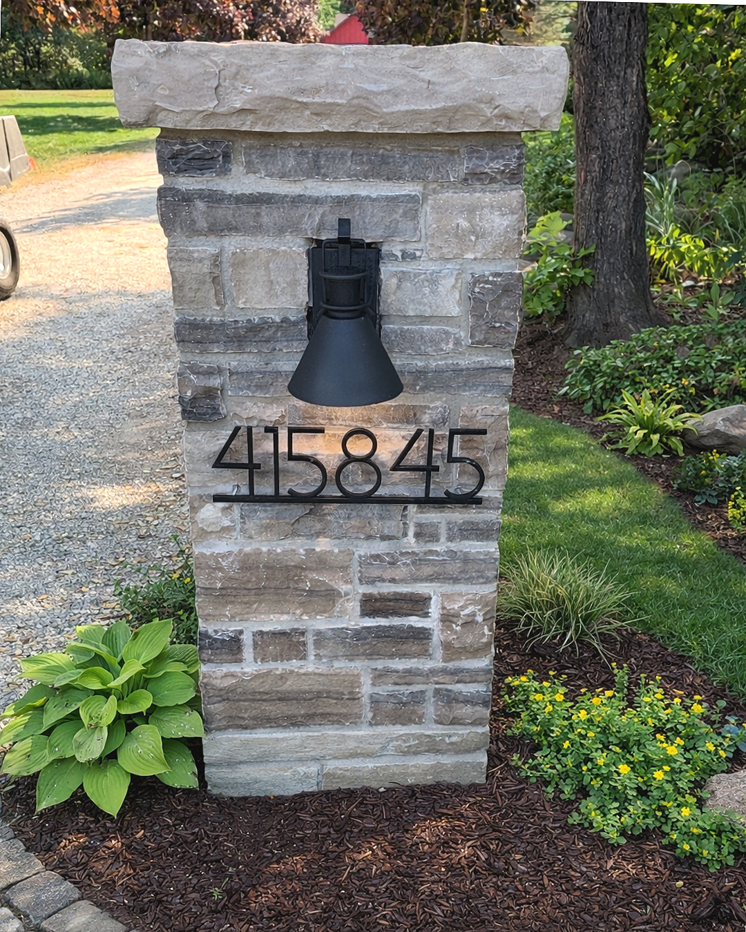 A stone mailbox with black house numbers 415845 and a black lamp attached to the front, surrounded by green plants, flowers, and a lawn.