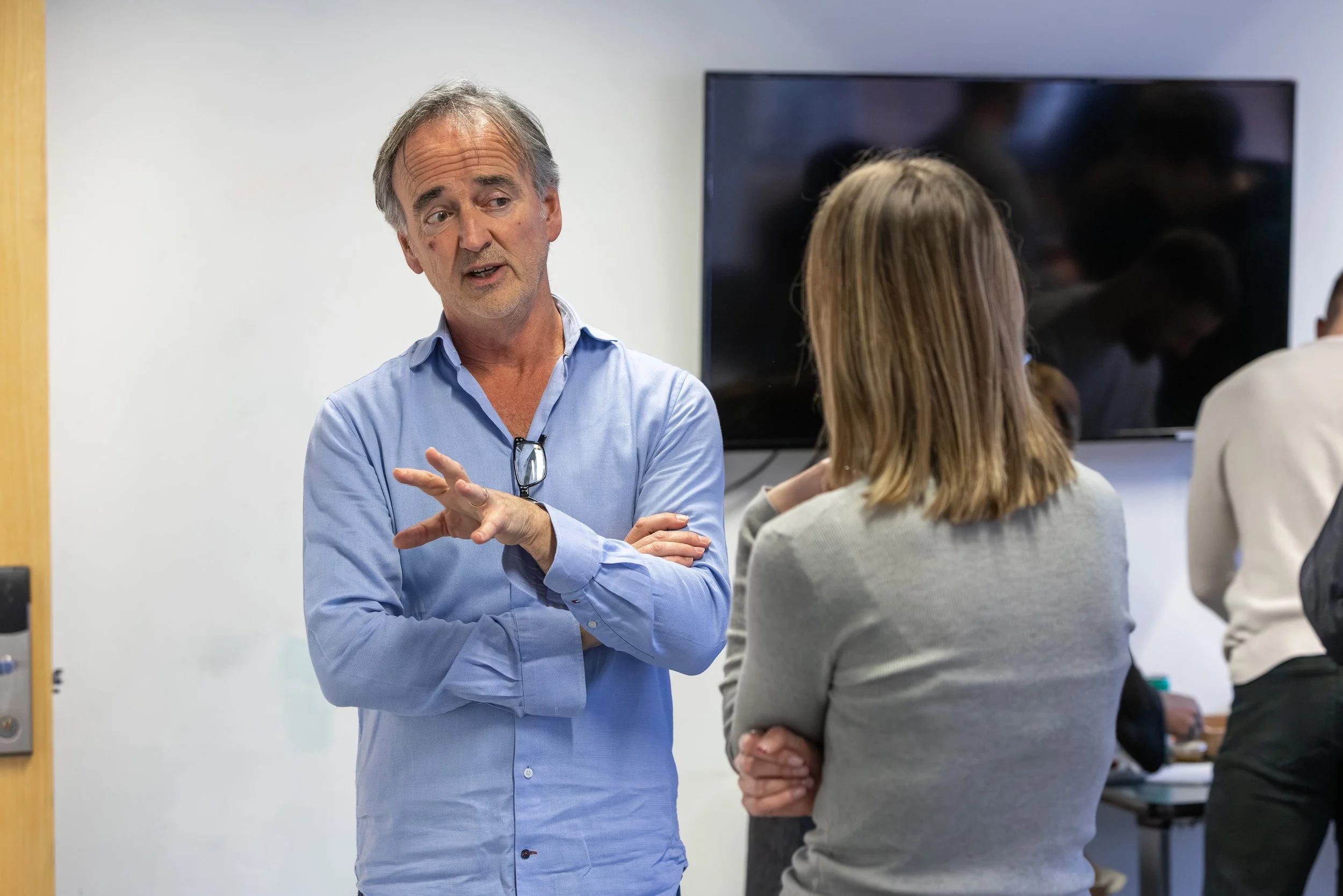 A man wearing a blue dress shirt and glasses hanging from his collar, talking to a woman with shoulder-length blonde hair in a gray sweater, in a room with a large TV screen on the wall behind them.