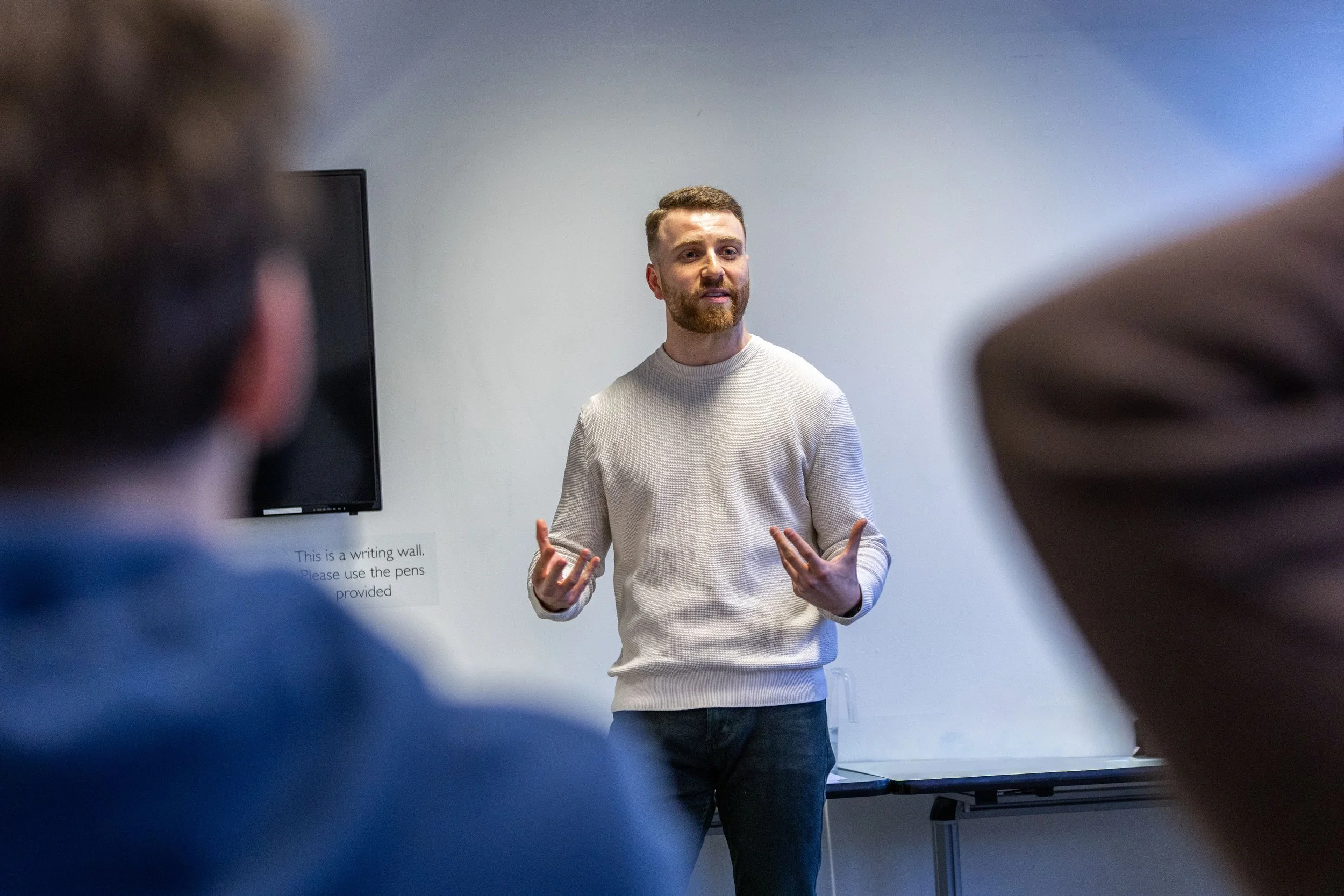 Man giving a presentation in front of a white wall with a TV screen, while speaking to an audience.