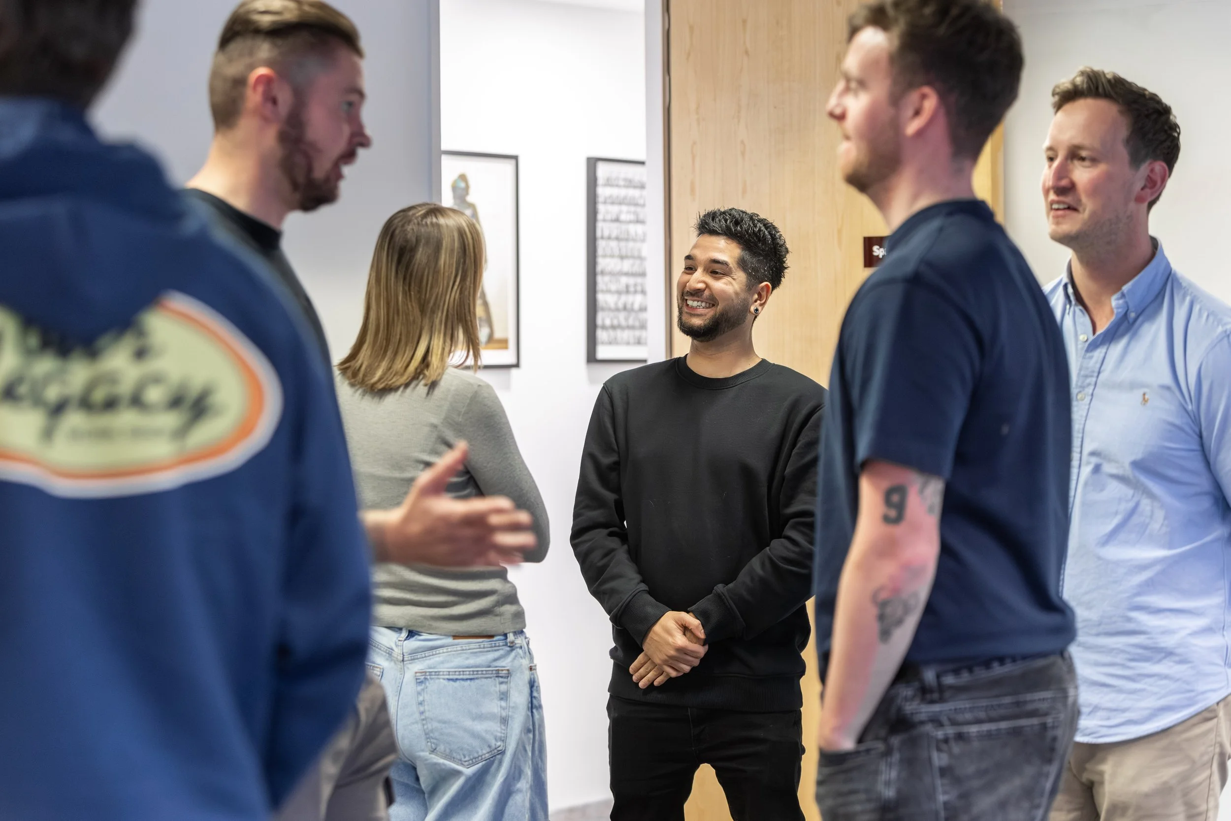 A group of five people socializing in an art gallery, smiling and engaging in conversation, with framed artwork on the walls in the background.