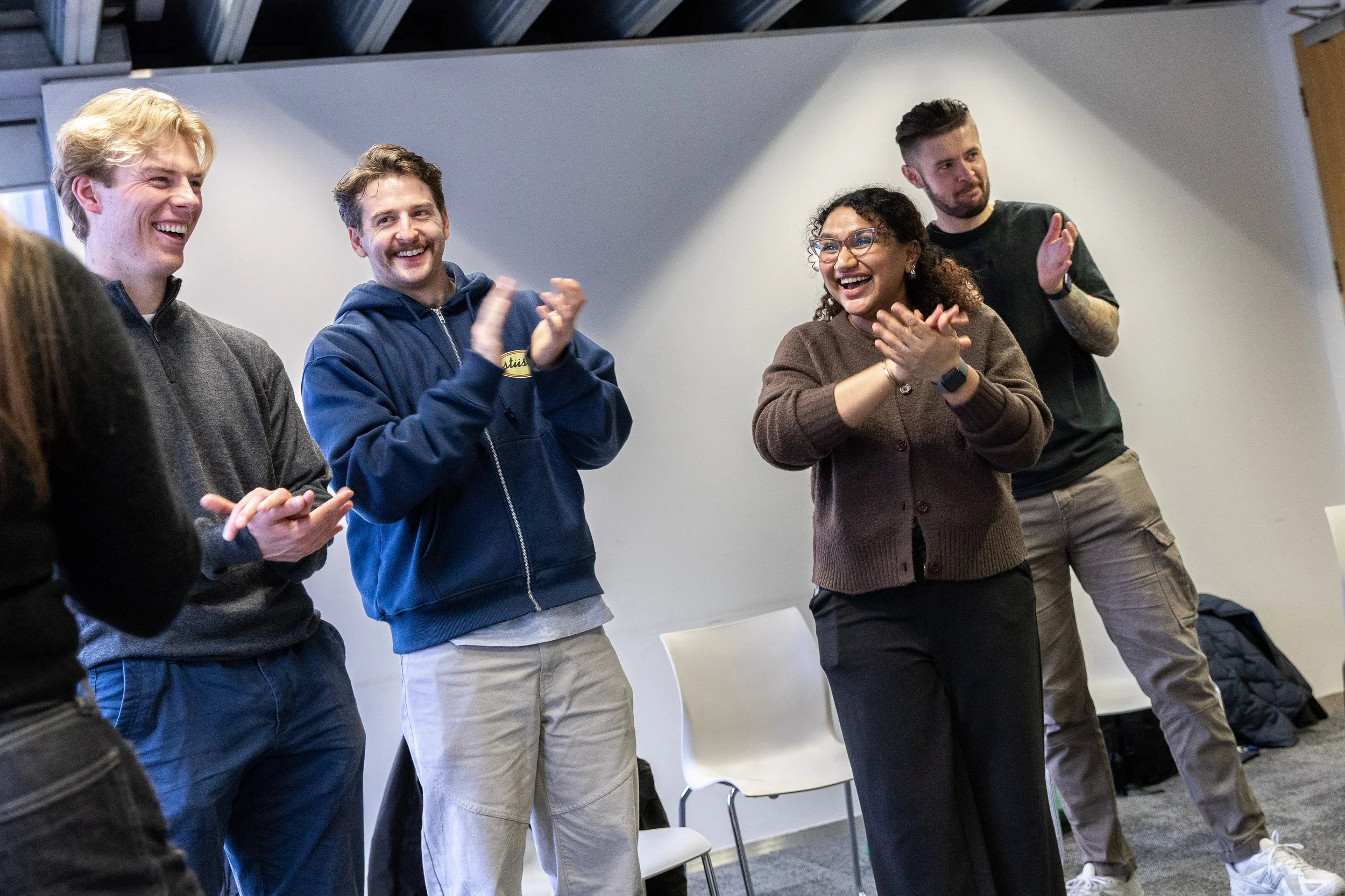 Group of five diverse young adults standing in a room, smiling and clapping, participating in an activity or celebration.
