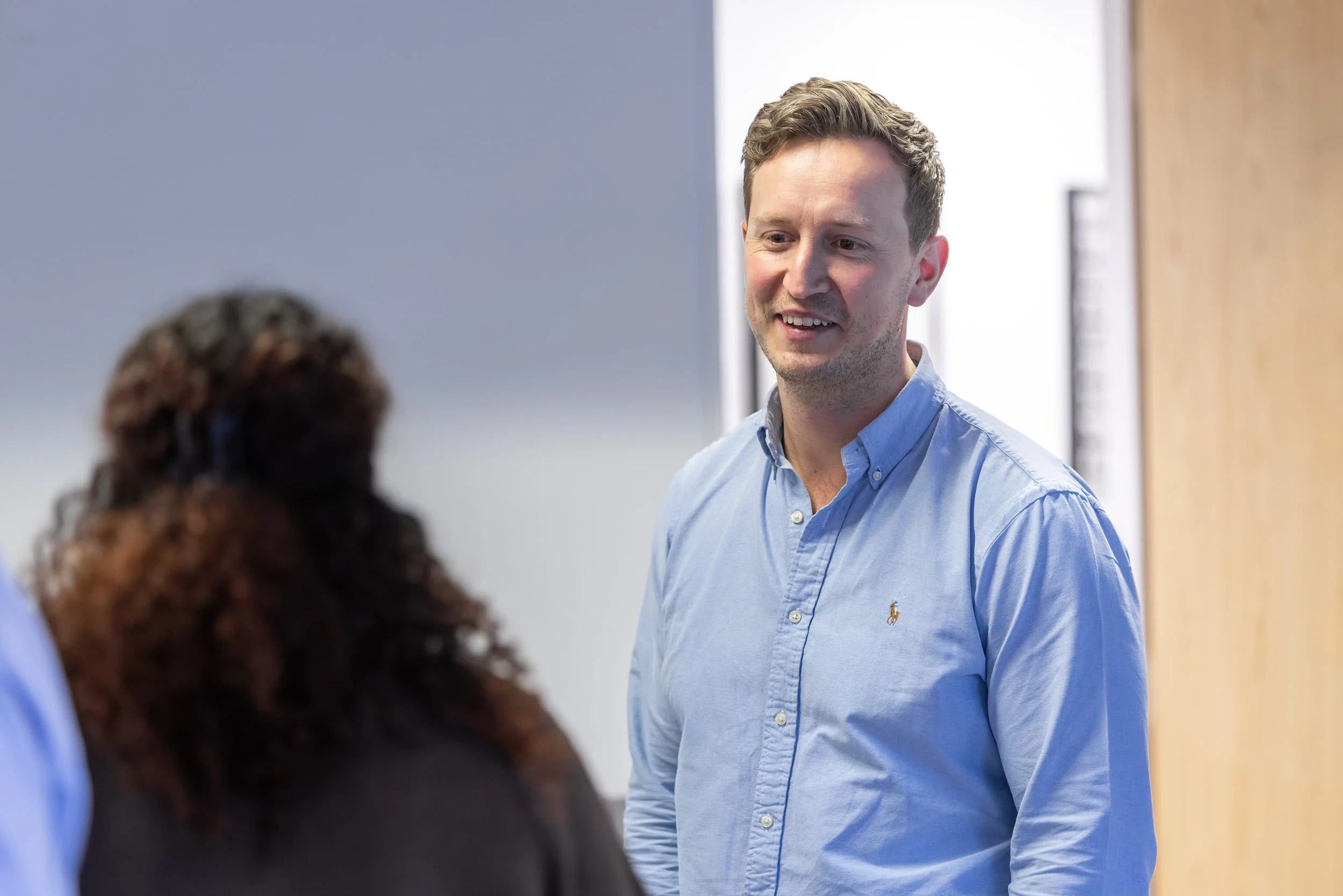 A man in a blue collared shirt smiling and talking to a woman with curly hair in a professional setting.