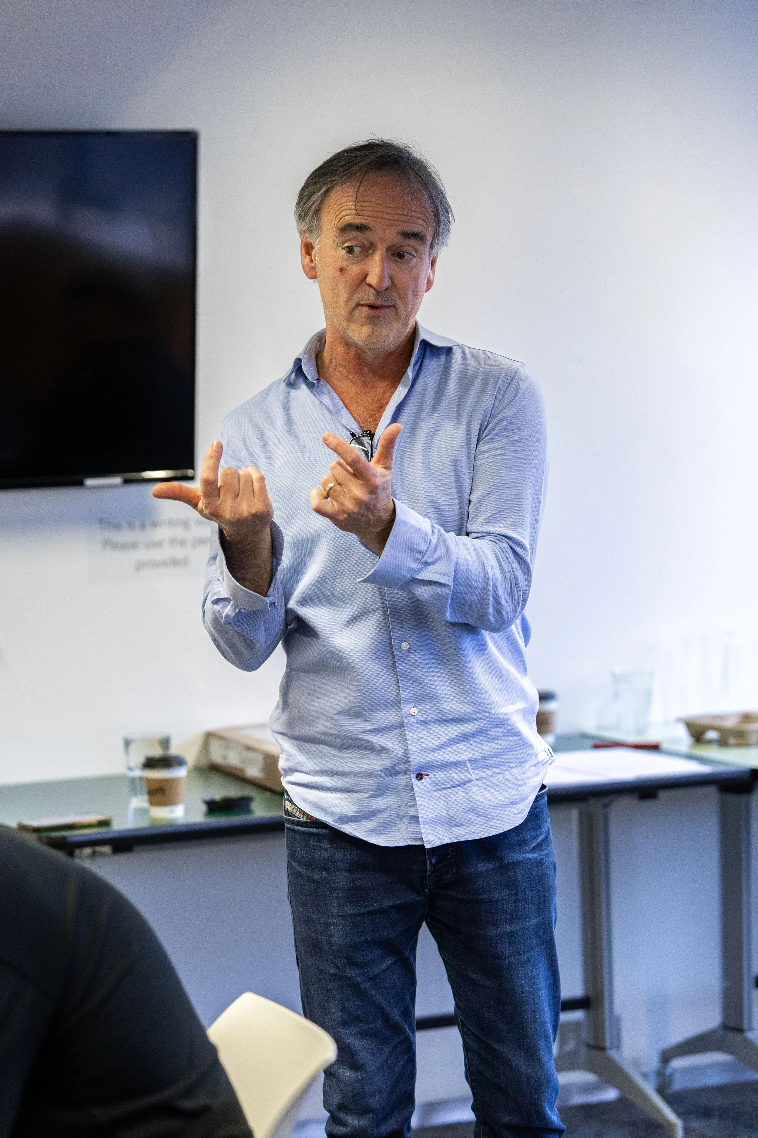 A man in a light blue shirt and jeans giving a presentation in a classroom or conference room.